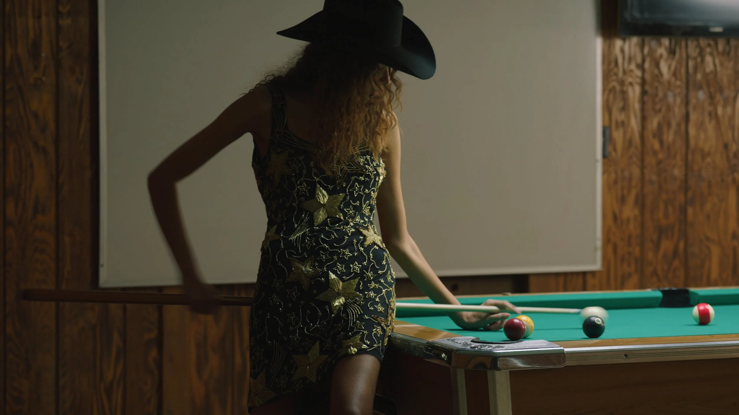 A woman with curly hair wearing a black cowboy hat and a black dress with gold star and butterfly embroidery, playing pool in a room with wood-paneled walls and a whiteboard in the background.