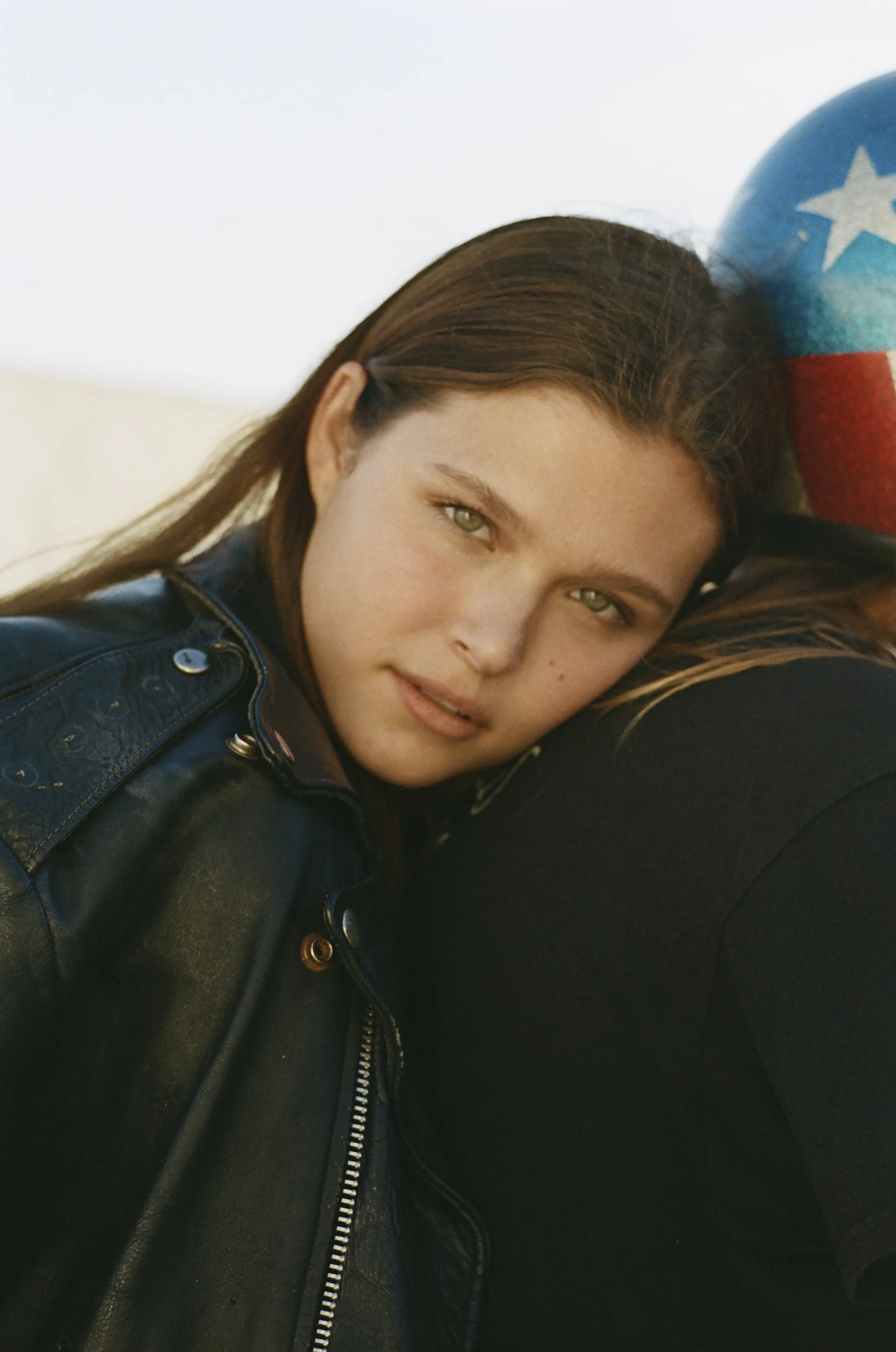A young girl with long brown hair and green eyes lying on a person's shoulder, partially visible, wearing a black leather jacket. The background is plain, and part of a patriotic-themed balloon with red, white, and blue colors and a star pattern is visible.