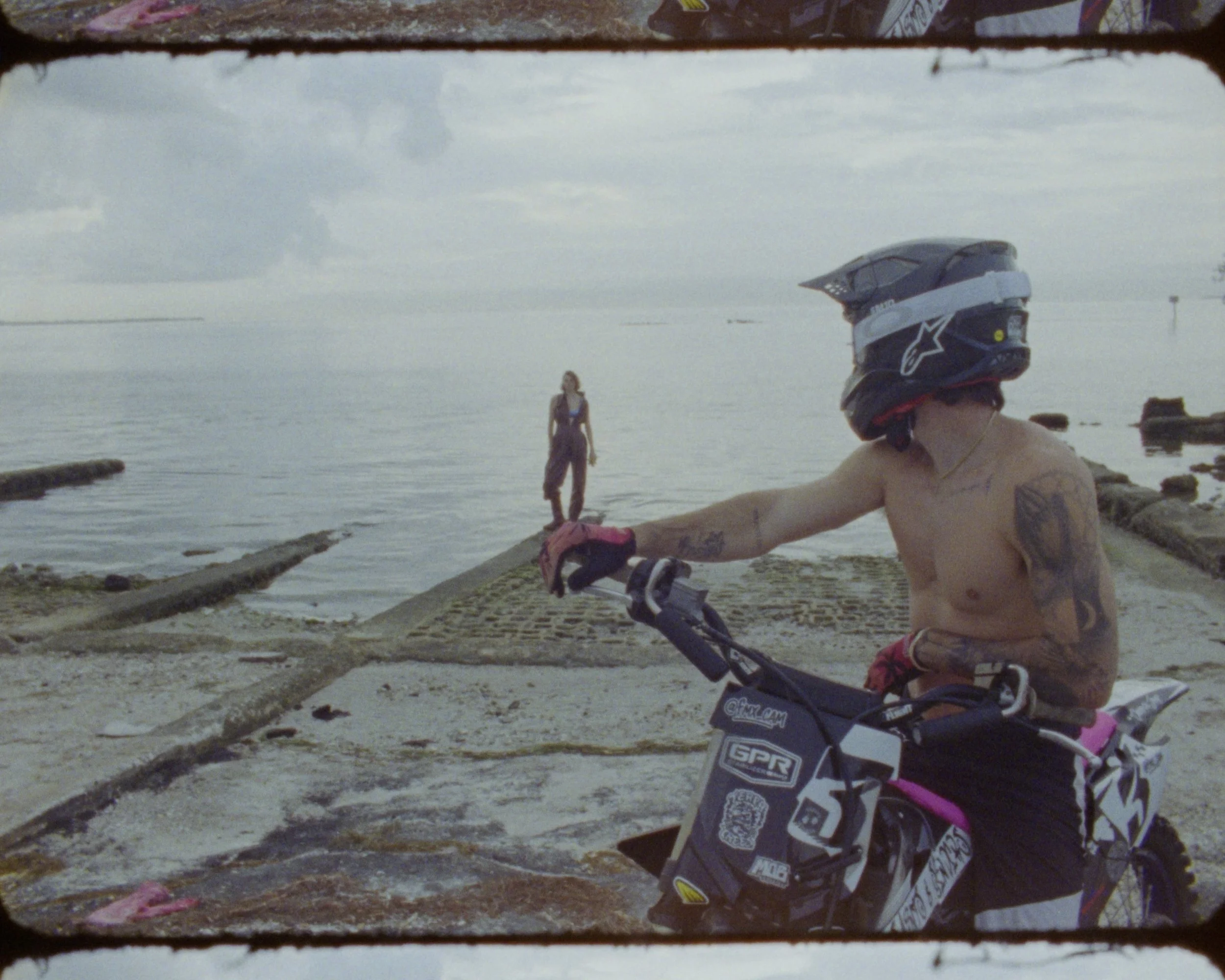 A shirtless man with a motorcycle helmet on his head riding a dirt bike on a beach. In the background, there is a woman standing near the water's edge and the ocean with a cloudy sky overhead.