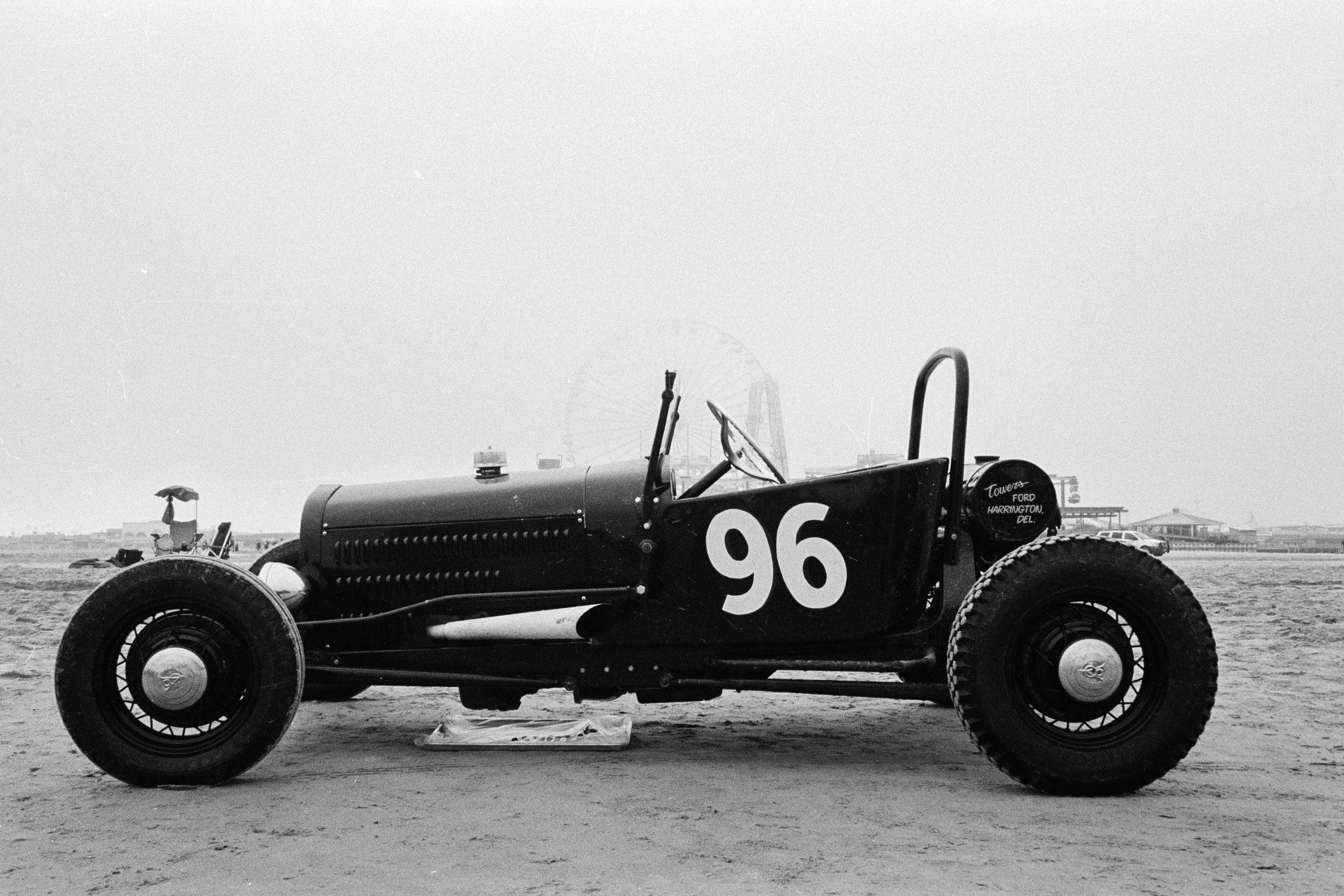 A vintage race car numbered 96 parked on a beach, with a person holding an umbrella and a wheelbarrow in the background, and a large Ferris wheel and buildings in the distance.