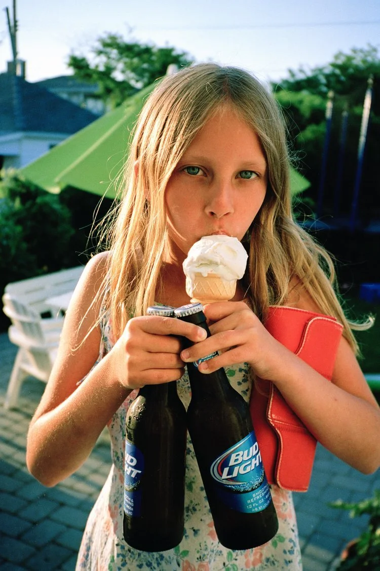 A girl with long blonde hair holding two Bud Light beer bottles in one hand and a vanilla ice cream cone in the other. She is outdoors on a patio with a green umbrella, trees, and houses in the background.