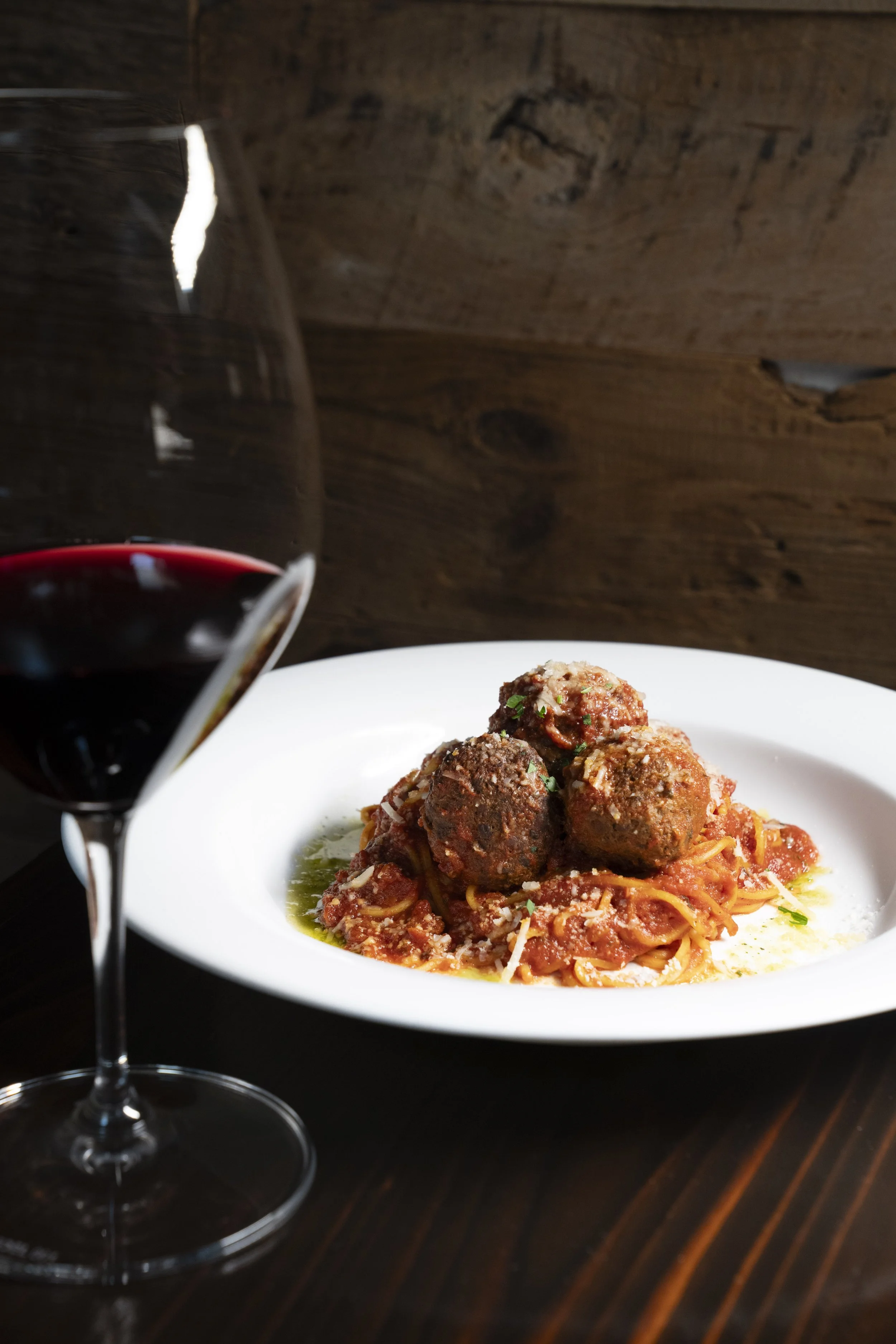 A plate of spaghetti with meatballs dressed in marinara sauce, garnished with grated cheese and herbs, next to a glass of red wine, on a dark wooden table.