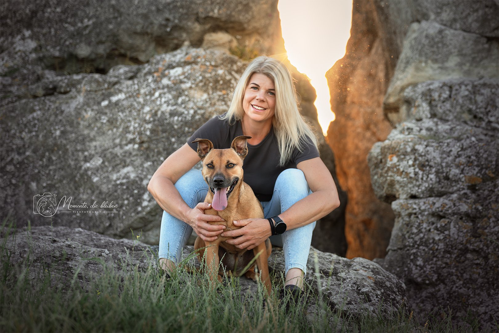Eine lachende Frau sitzt mit einem braunen Hund zwischen Felsen während des Sonnenuntergangs im Freien.