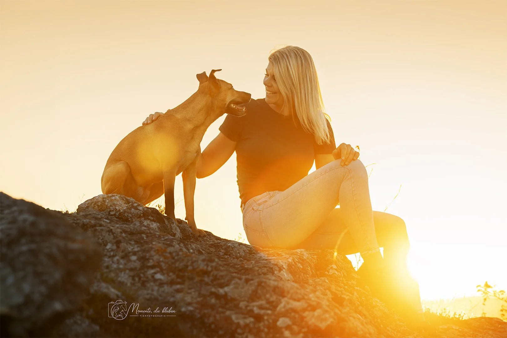 Frau und Hund sitzen auf einem Felsen bei Sonnenuntergang, lächeln sich an.