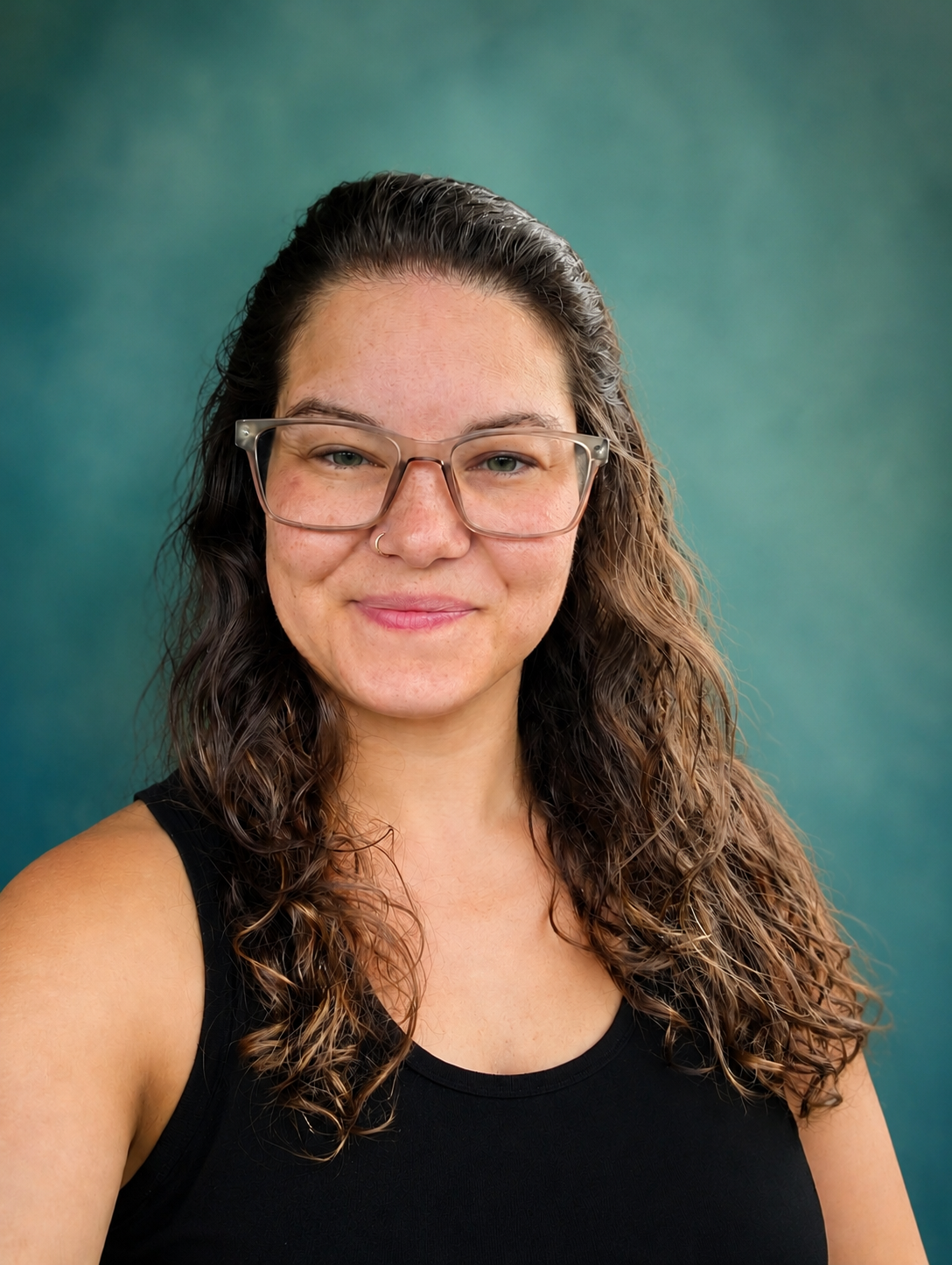 A young woman with wavy brown hair, wearing glasses and a black tank top, smiling in front of a blurred green background.