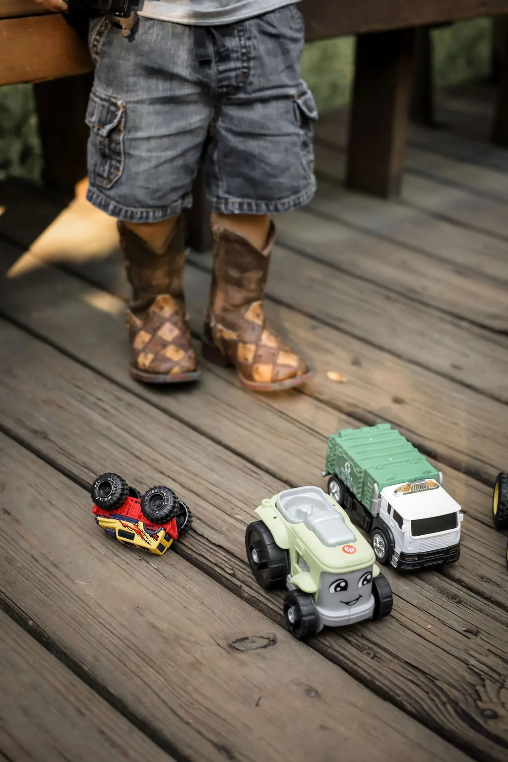 A child's legs wearing camo boots, denim shorts, and a white shirt, standing on a wooden deck next to small toy cars and trucks on the ground.
