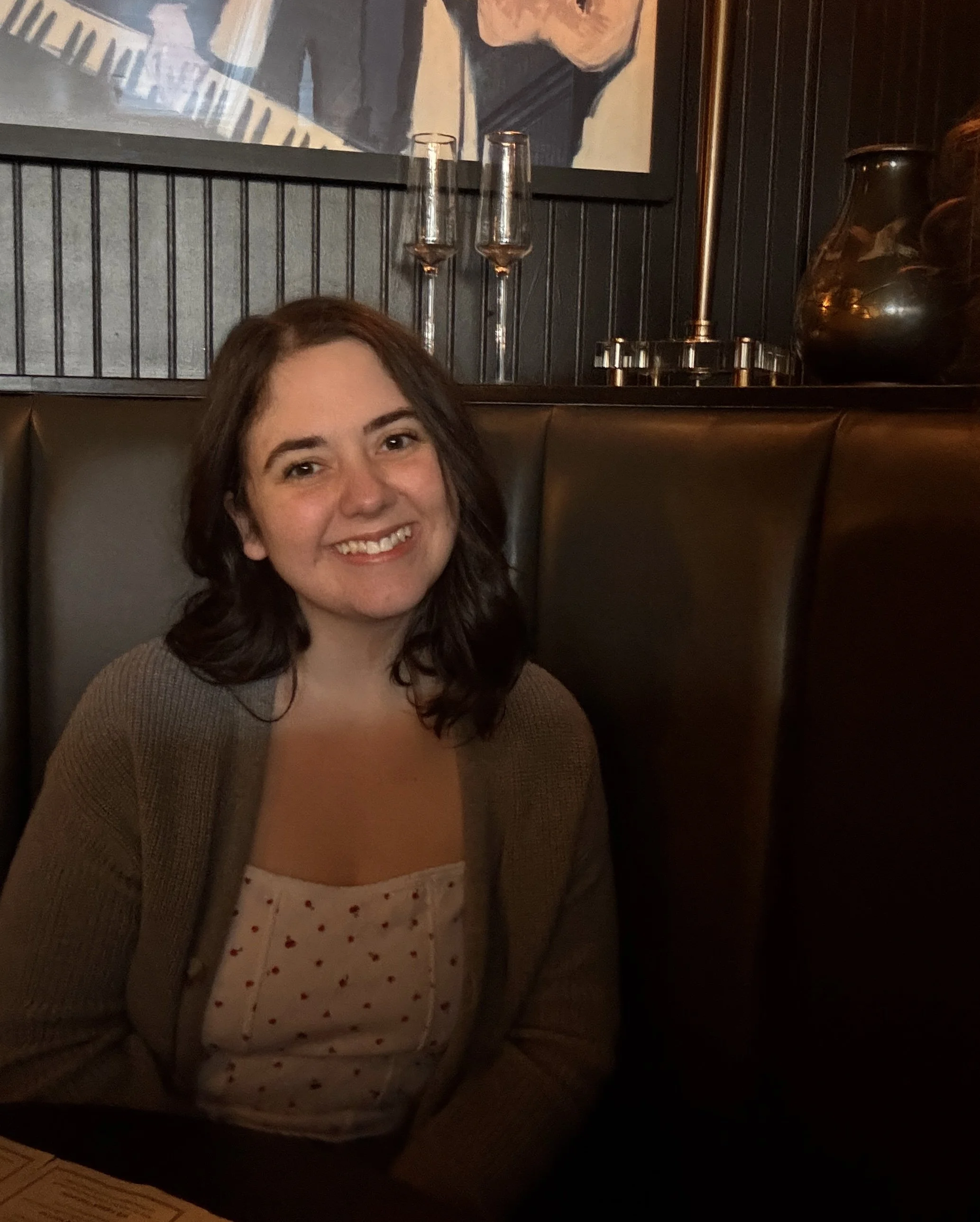 A woman with shoulder-length dark hair smiling and sitting against a dark wall with framed artwork and decorative vases behind her. She is wearing a beige cardigan and a white top with red hearts.
