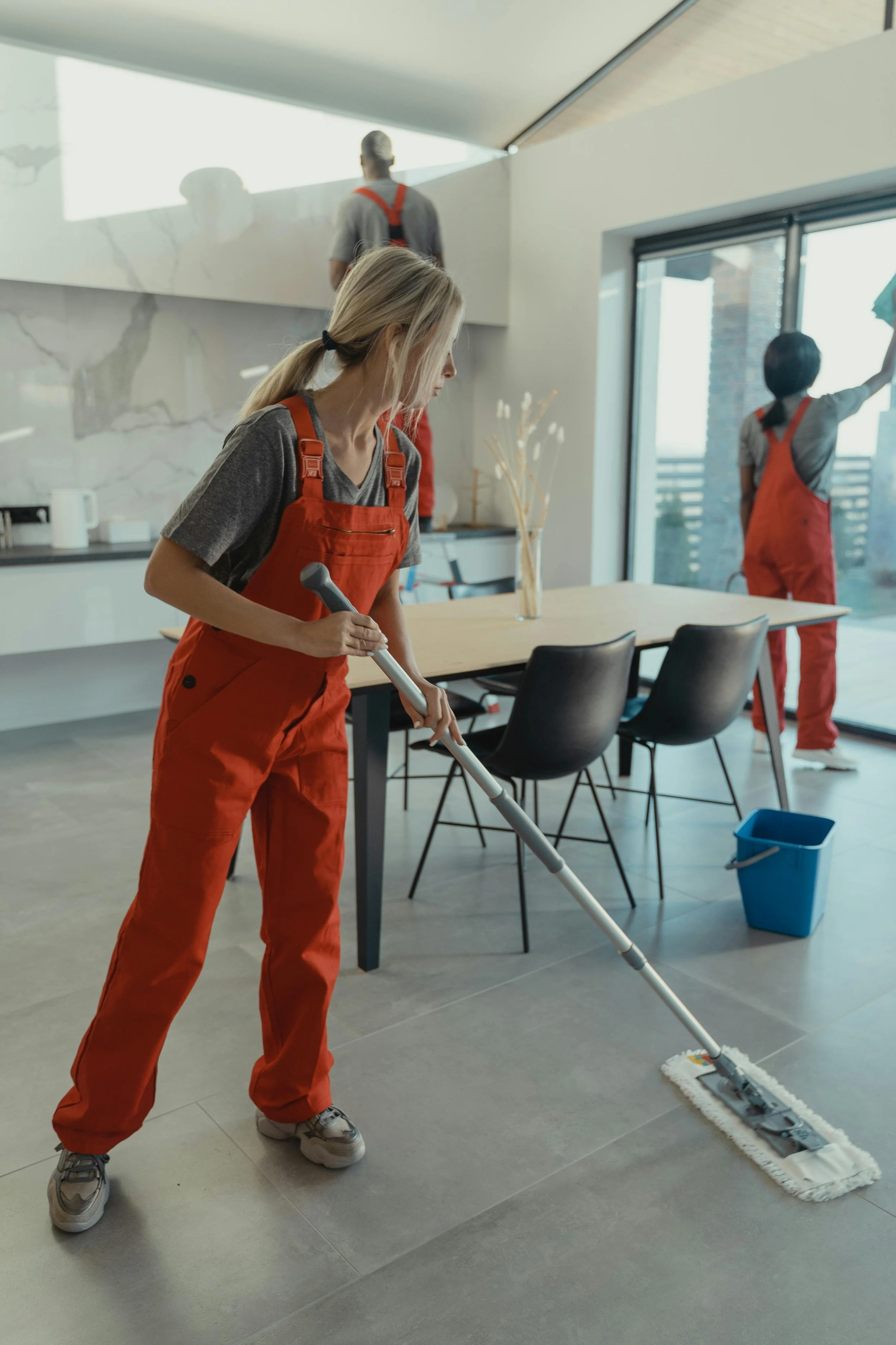 A woman wearing orange overalls and sneakers is mopping the floor in a modern kitchen or dining area, with a blue mop bucket nearby. Two other people in similar overalls are in the background, one cleaning a window and the other standing on a ledge.