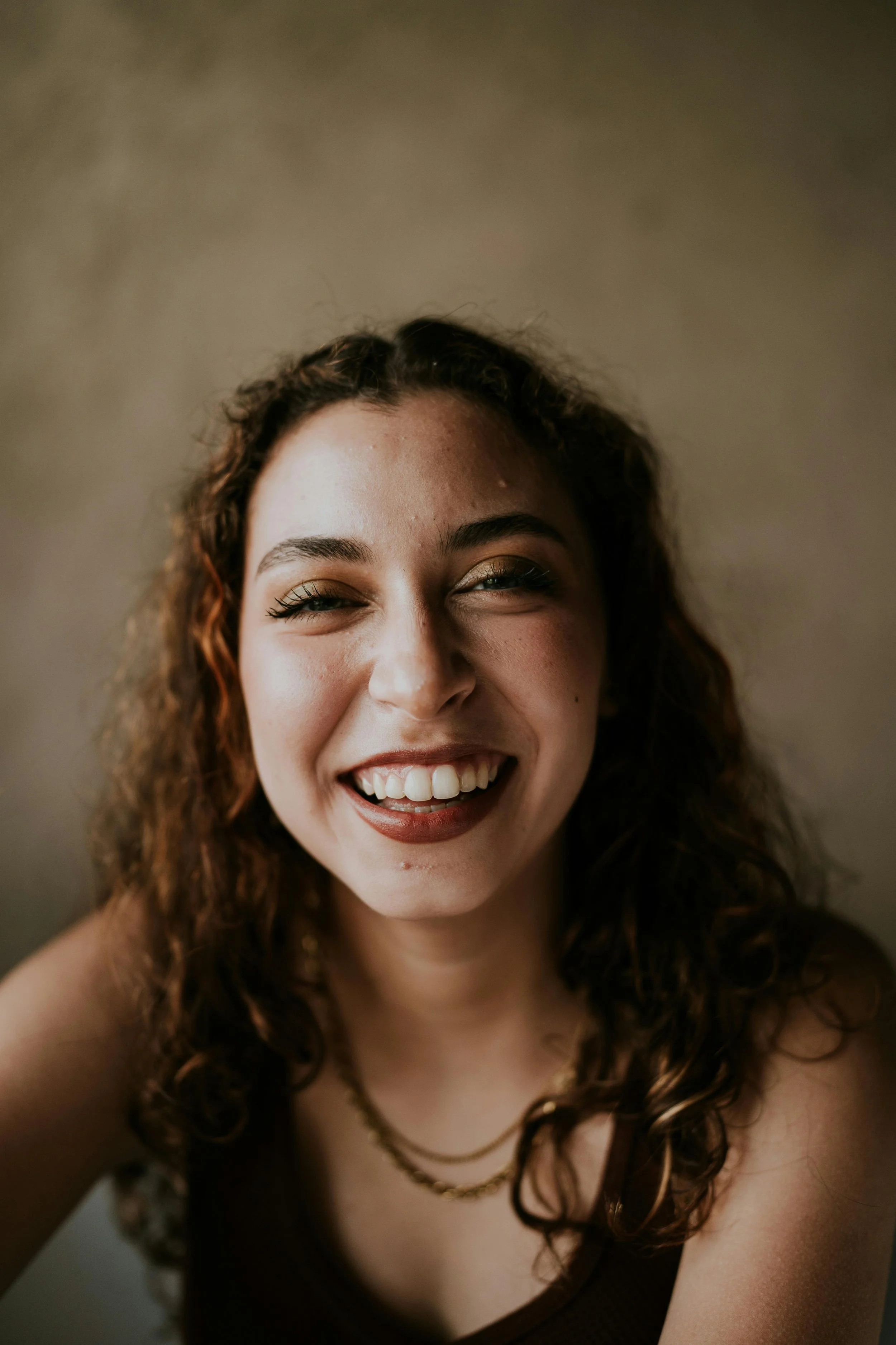 Close-up of a woman with curly hair smiling, showing her teeth, wearing eye makeup and a black top with layered necklaces.