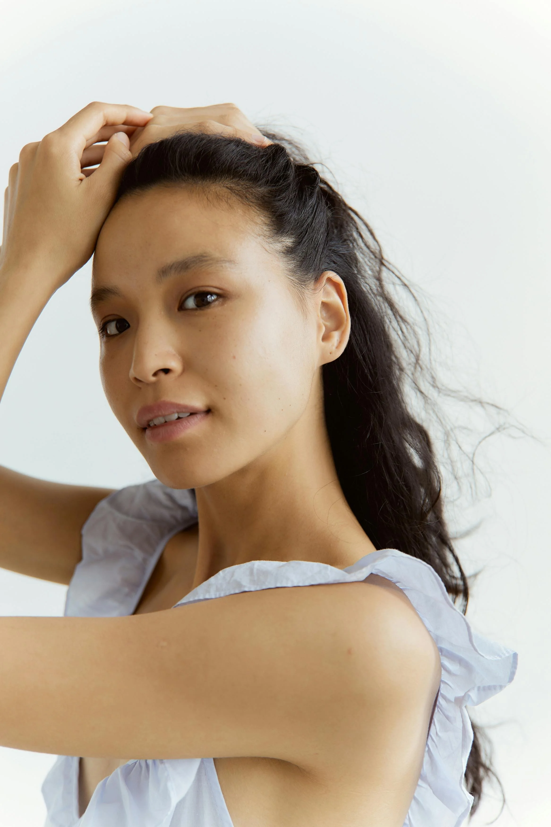 A woman with long dark hair in a ponytail, wearing a white ruffled top, looks at the camera while holding her head with one hand, against a plain white background.