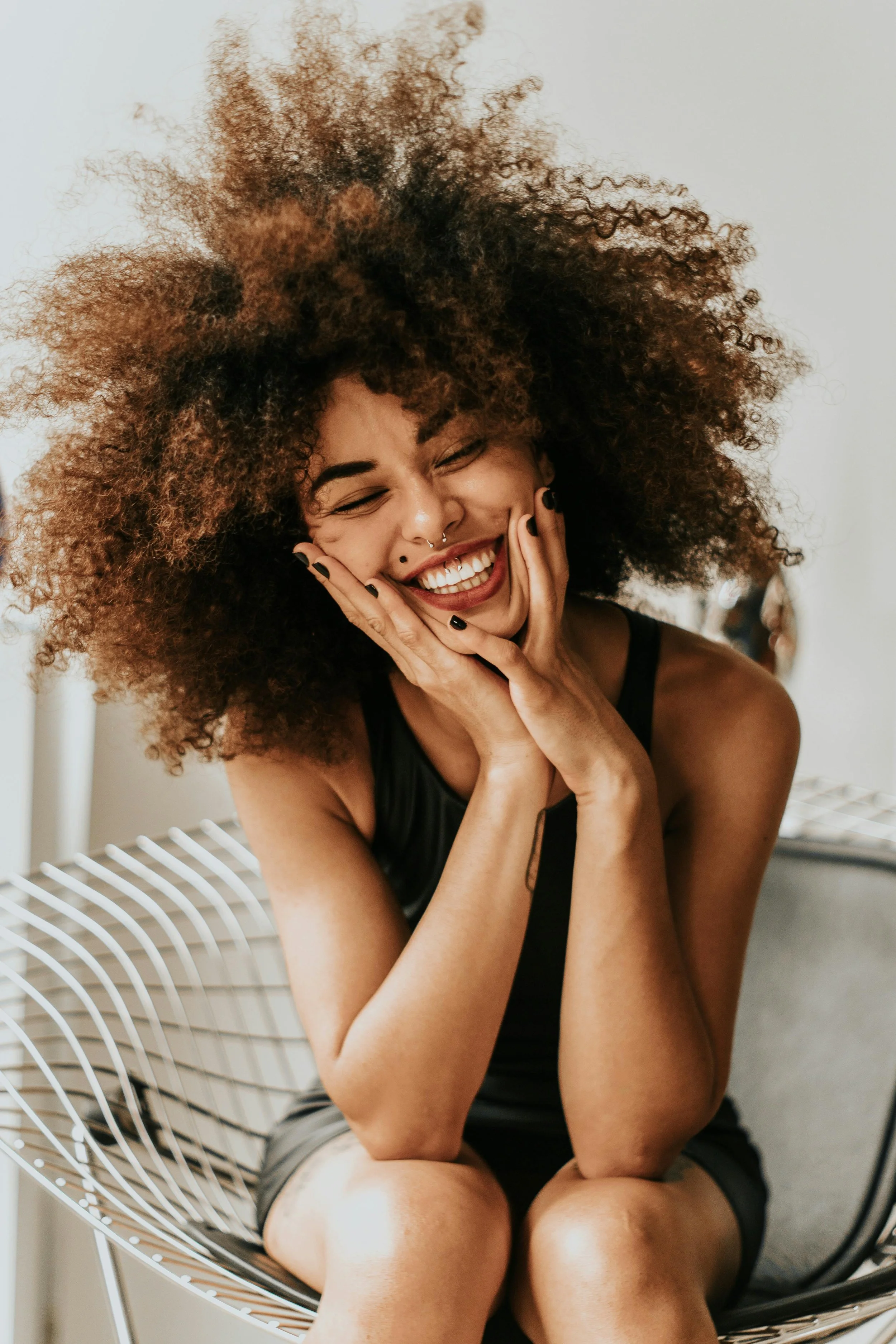 A woman with curly hair sitting on a white wire chair, smiling with eyes closed, touching her face with both hands.
