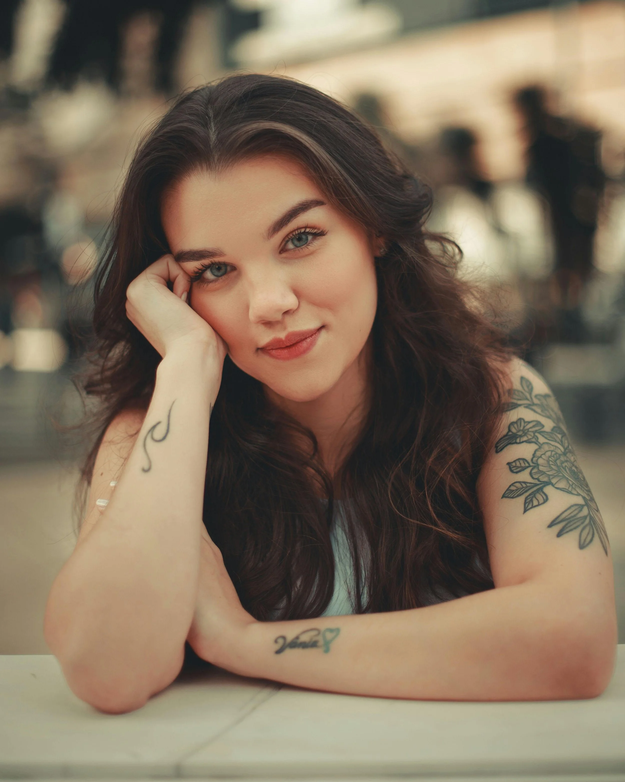 Close-up of a smiling woman with long dark hair, blue eyes, and tattoos on her arm, resting her head on her hand.