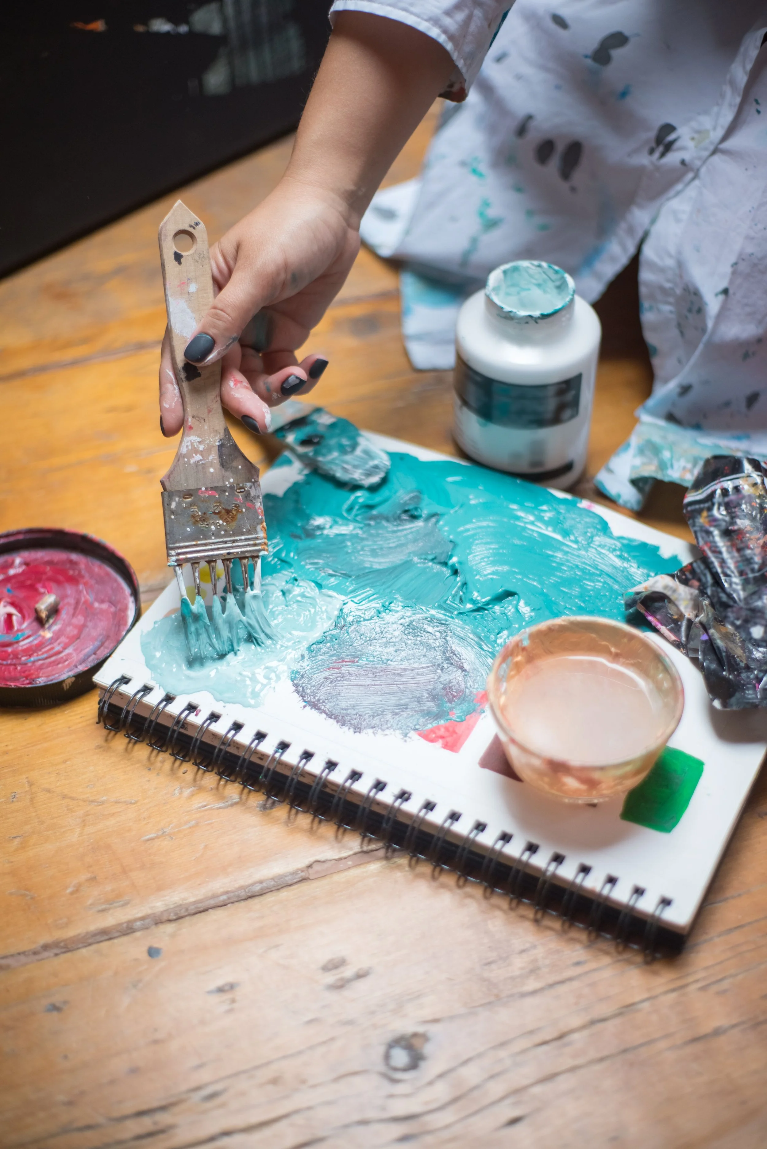 A person mixing blue paint on a sketchbook with a wide paintbrush, surrounded by open paint containers and splattered paint on their white shirt and the wooden table.