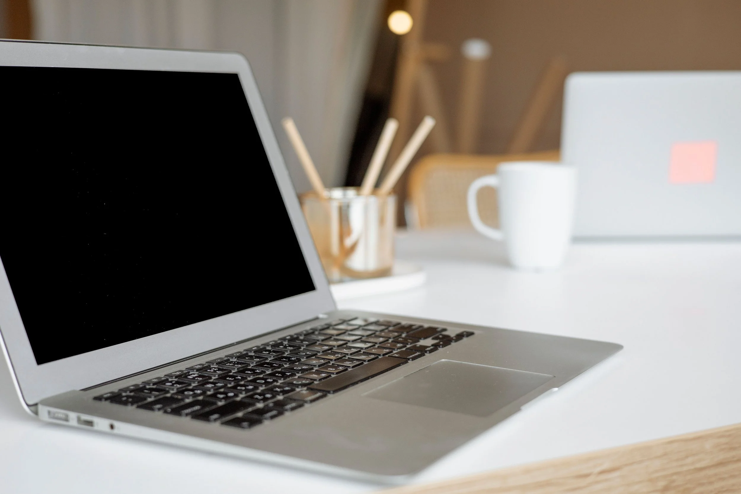 A close-up of a silver laptop on a white desk with a black screen, with a white mug and a glass jar with wooden stirring sticks in the background.