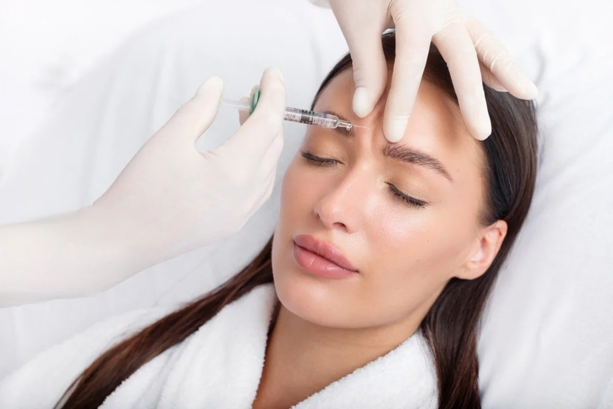 A woman receiving a Botox injection in her forehead while lying down with her eyes closed, with medical gloves and syringe visible.