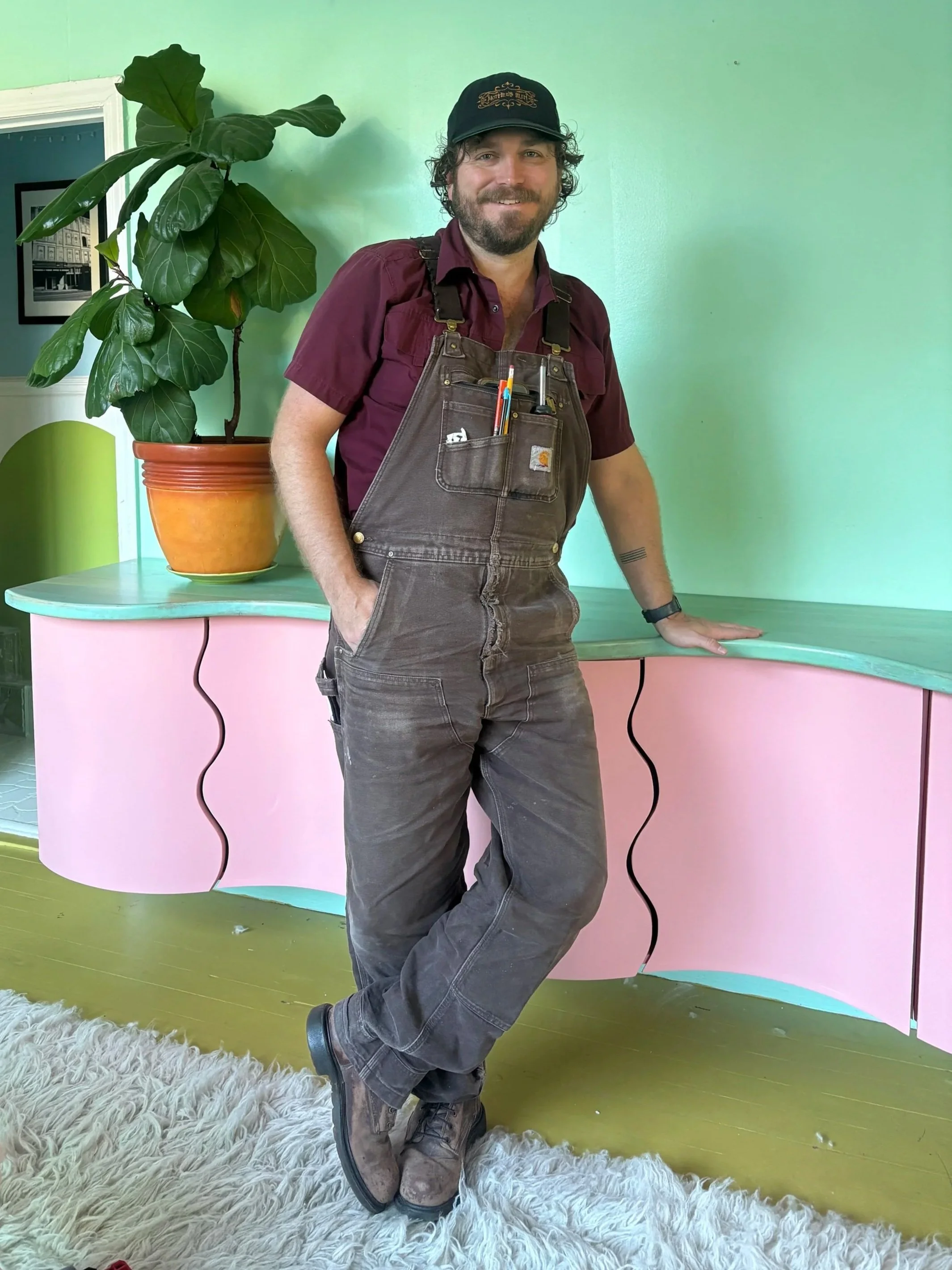 A man with curly hair and a beard, wearing a black cap, maroon shirt, and brown overalls, stands relaxed with one leg crossed over the other, leaning against a pink counter with a green top. Behind him is a large potted plant with broad, dark green leaves, and next to the plant is a black and white framed photo on the wall. The floor is green, and there is a white shaggy rug in front of him.