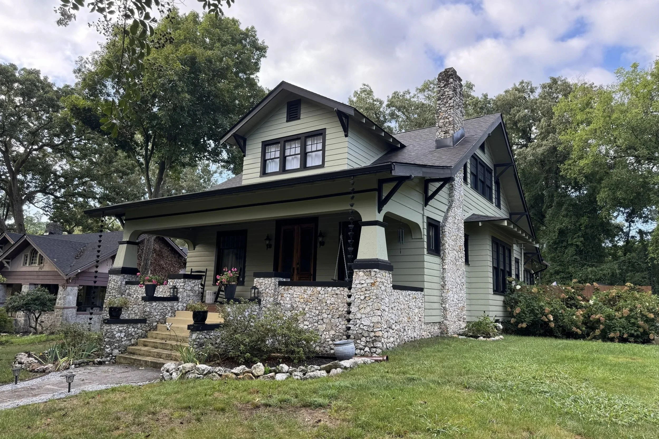 A house with light green exterior, black trim, stone chimney, front porch, surrounded by trees, with a lawn and garden.