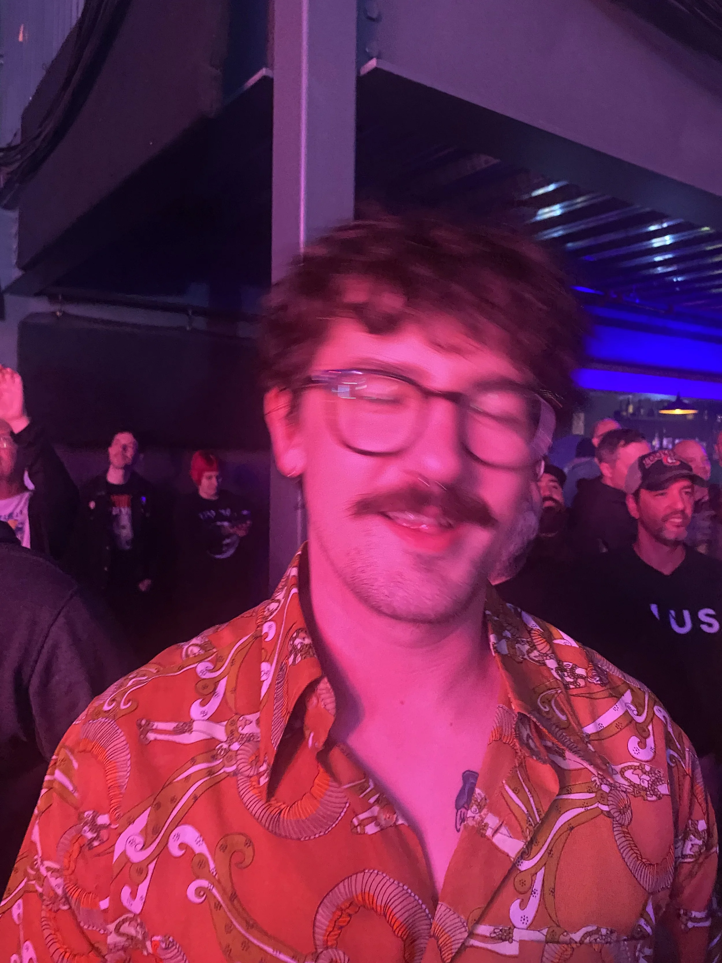 A smiling man with glasses and a mustache, wearing a vibrant orange shirt with a dragon and snake pattern, at a social event with purple and blue lighting.