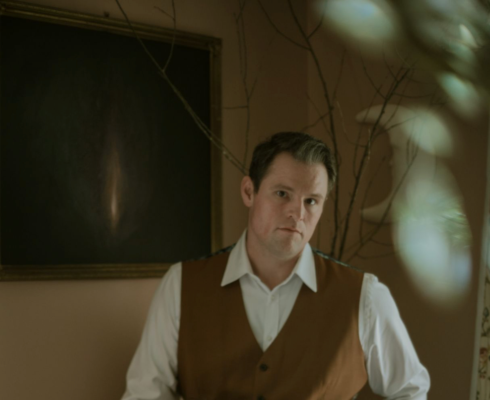 A man with dark hair and a serious expression, wearing a white shirt and brown vest, standing indoors with a chalkboard, branches, and blurred lights in the background.