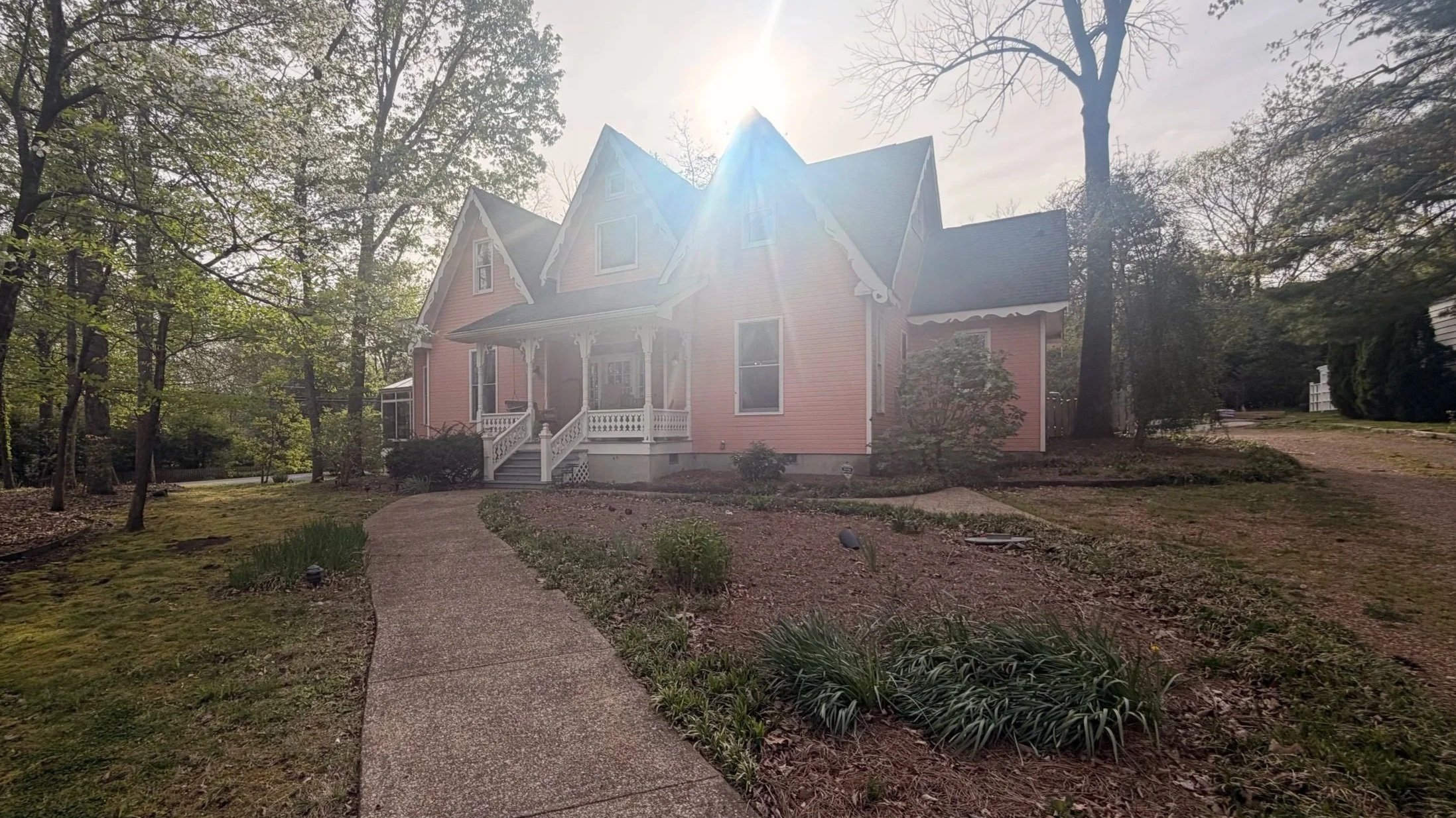 A pink Victorian style house with a front porch, surrounded by a garden with a curved pathway, trees, and bushes. The sun is shining brightly behind the house.