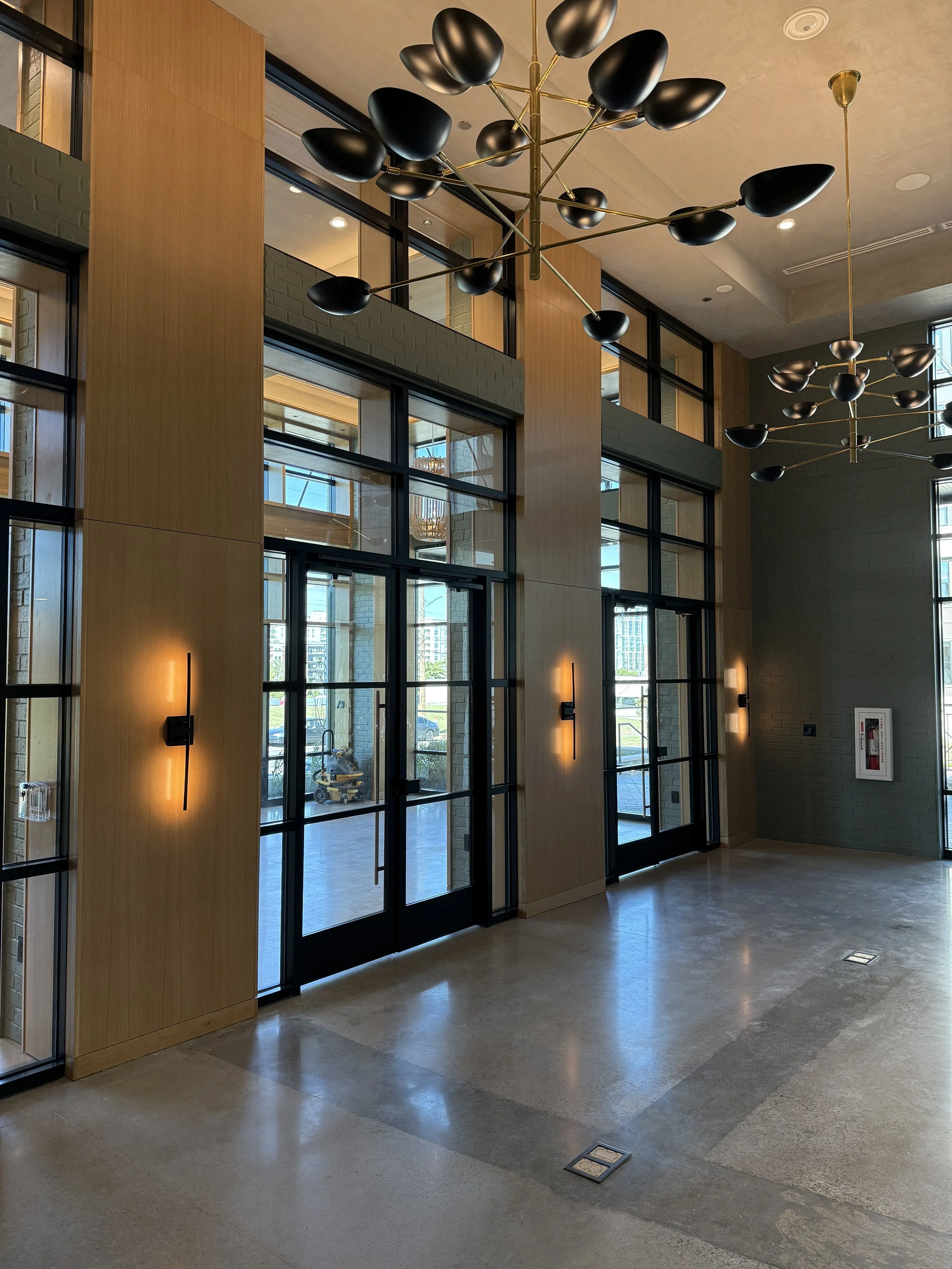 Modern interior lobby area with large glass doors, wooden walls, contemporary black and gold chandeliers, wall lights, and a polished concrete floor.