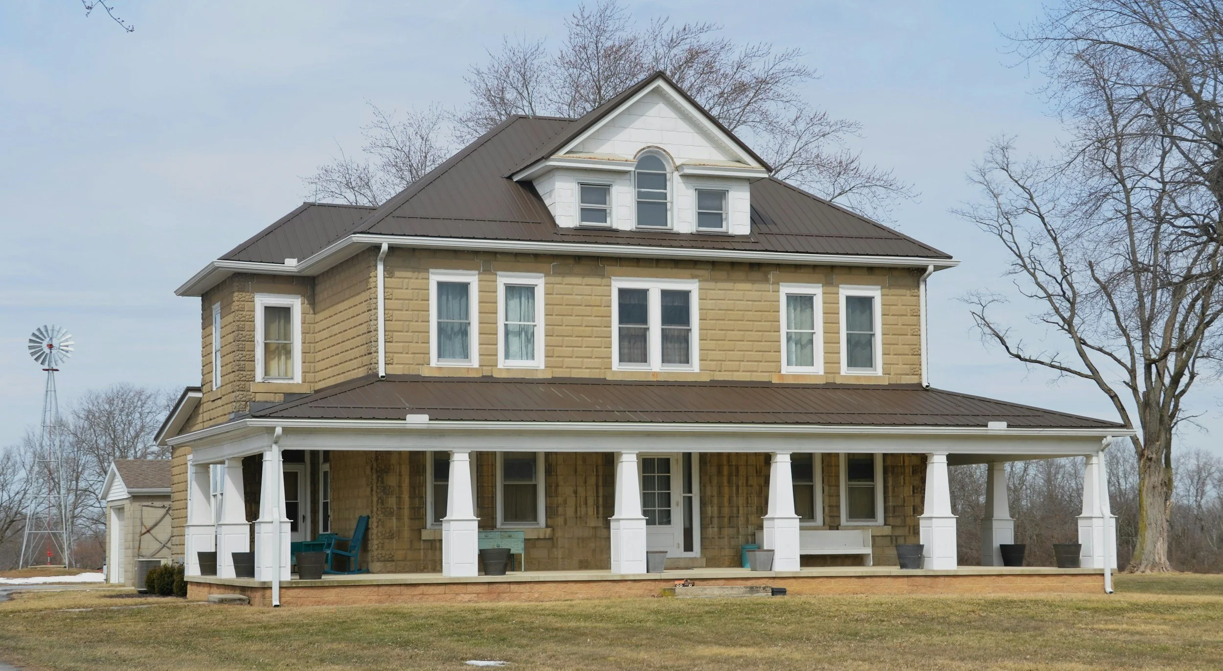 A two-story house with beige brick walls, white trim, and a dark metal roof. The front porch has white columns, chairs, and plants, with leafless trees and a windmill in the background.