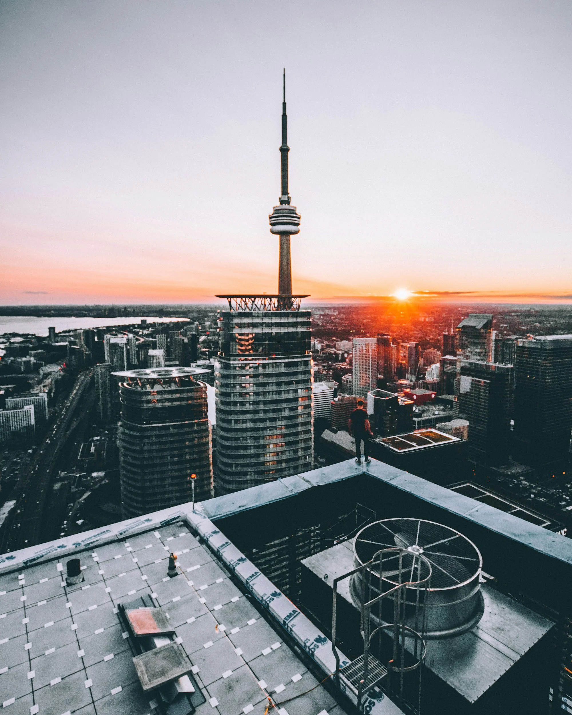 View from a rooftop of Toronto's CN Tower during sunset, with the city skyline and buildings in the foreground and the sunset on the horizon.