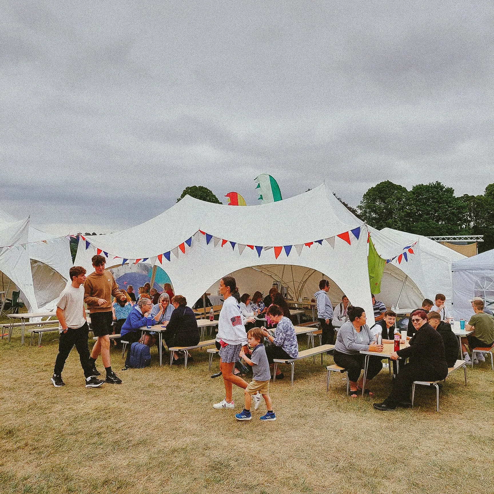 Crowd of people gathered under a large white tent at an outdoor event, with some sitting at tables and others walking around. Banners in red, white, and blue decorate the tent, and colorful flags are attached to the tent's top. Overcast sky and trees in the background.