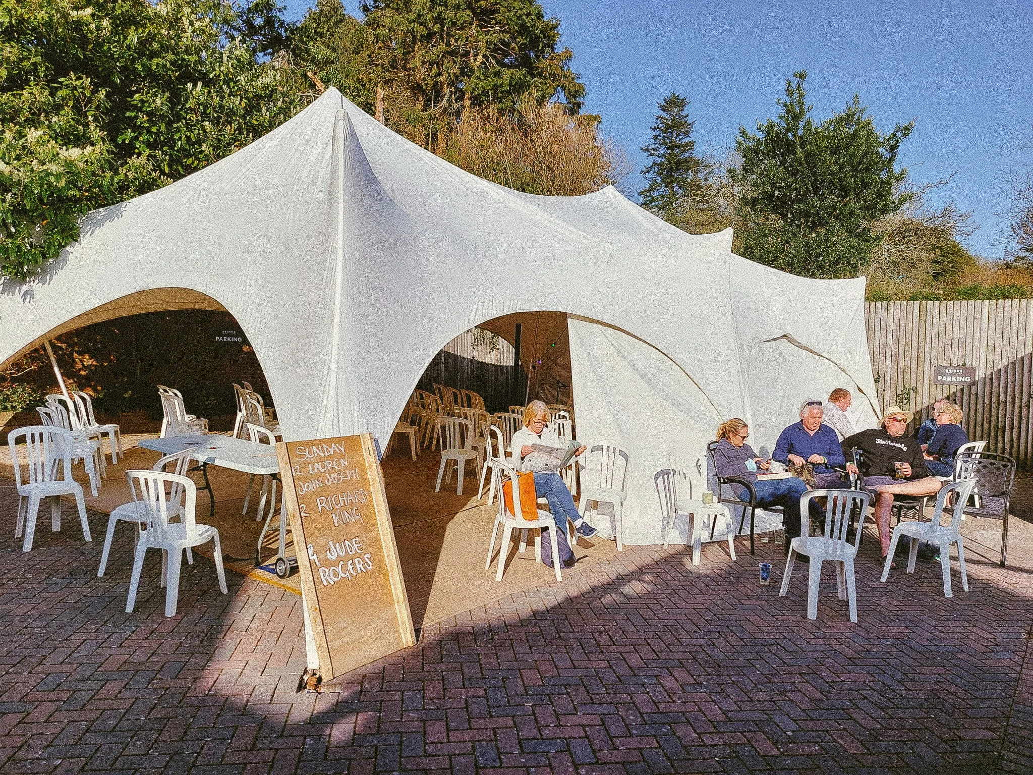 Outdoor gathering under a white tent with people sitting and chatting, surrounded by trees and a wooden fence on a sunny day.