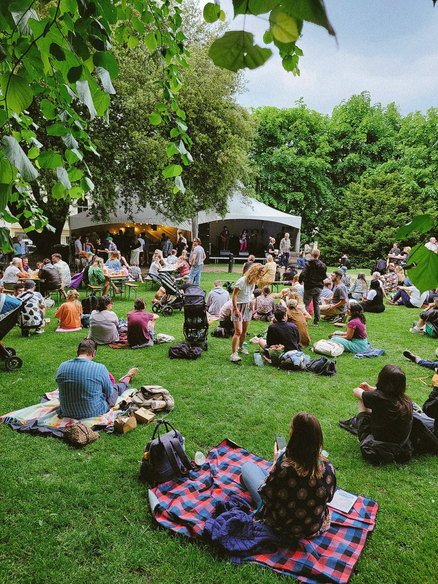 A large outdoor gathering at a park or garden with many people sitting on the grass, some on blankets, others at tables, with a stage and band performing under a tent. Trees surround the area, creating a shaded environment.