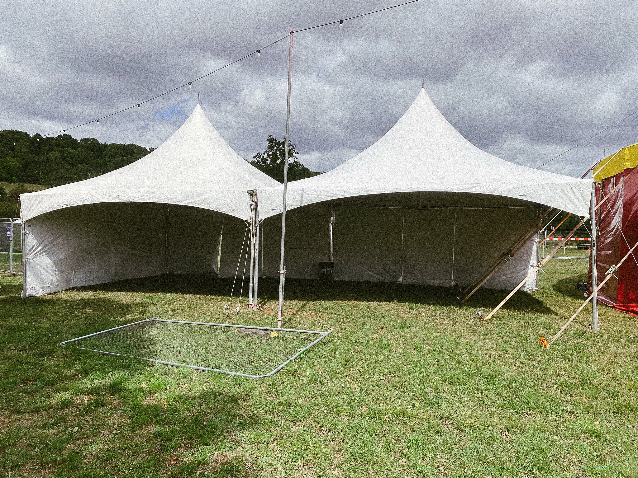Two white event tents set up on a grassy field under a cloudy sky, with string lights hanging above.
