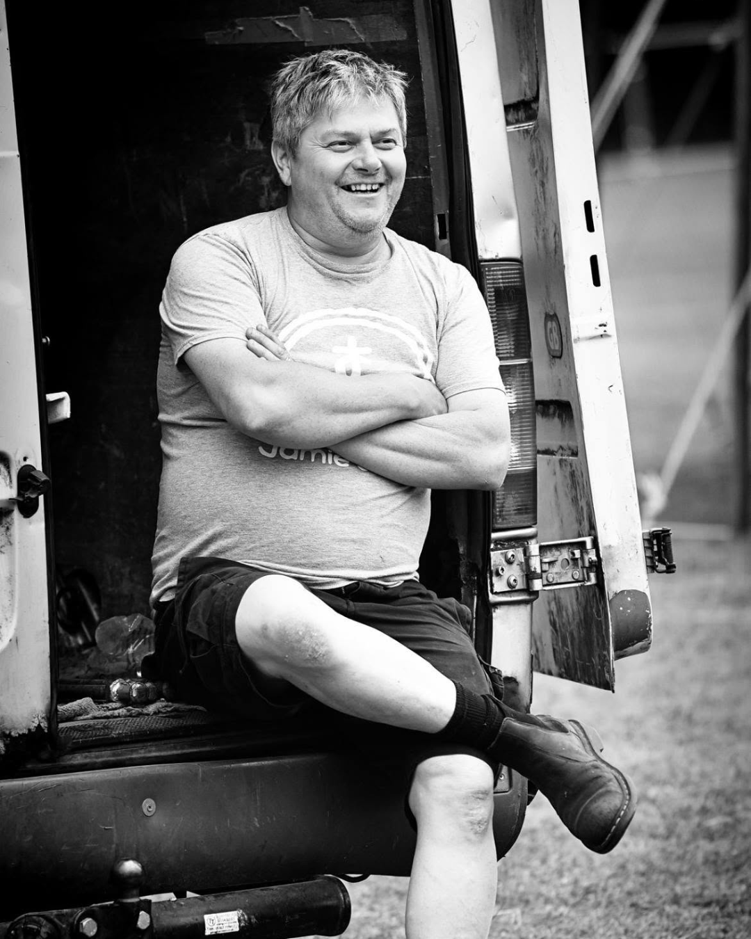 A man with curly hair and a beard smiling while sitting cross-legged at the back of a utility vehicle, dressed in a t-shirt, shorts, and work boots.