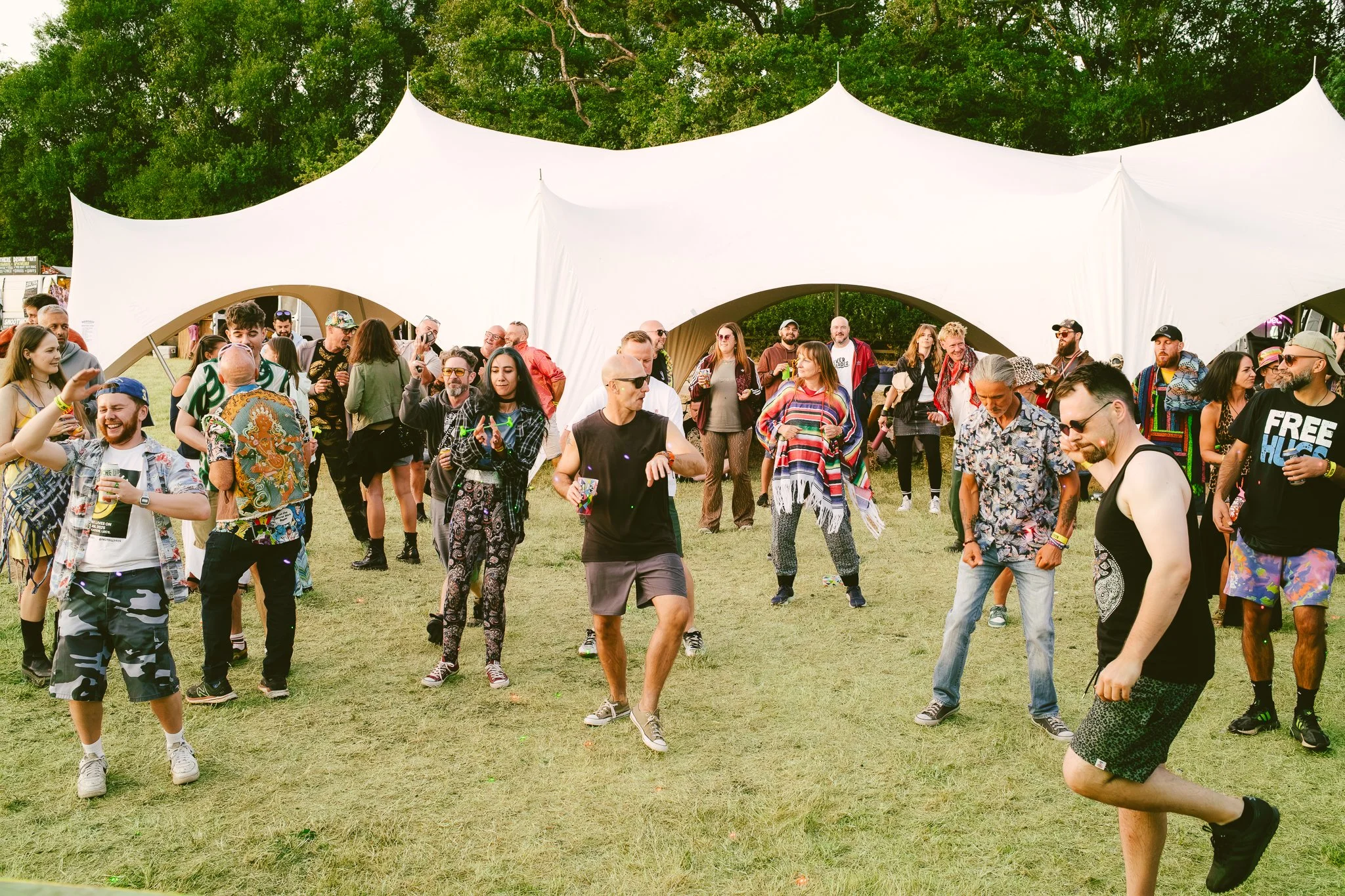 Crowd of people dancing and socializing outdoors under a large white tent at a festival, with trees in the background.