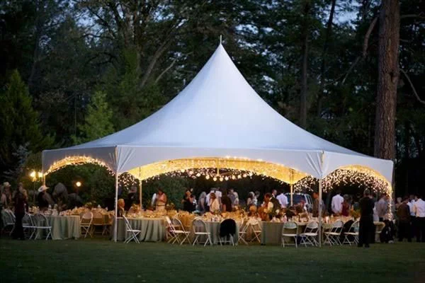 Outdoor wedding reception under a large white tent with string lights, tables, chairs, and guests, set in a wooded area during dusk.