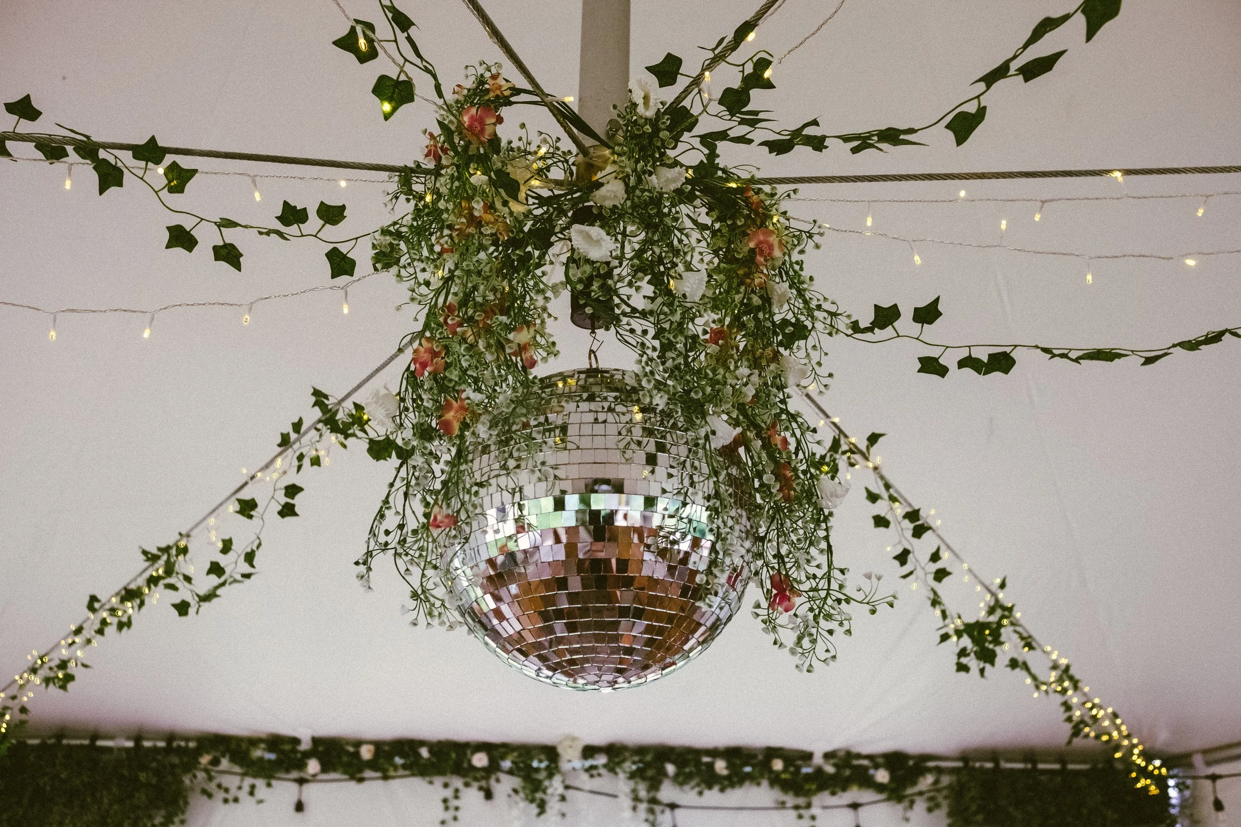 A disco ball chandelier decorated with green ivy, pink and white flowers, and small string lights hanging from the ceiling.