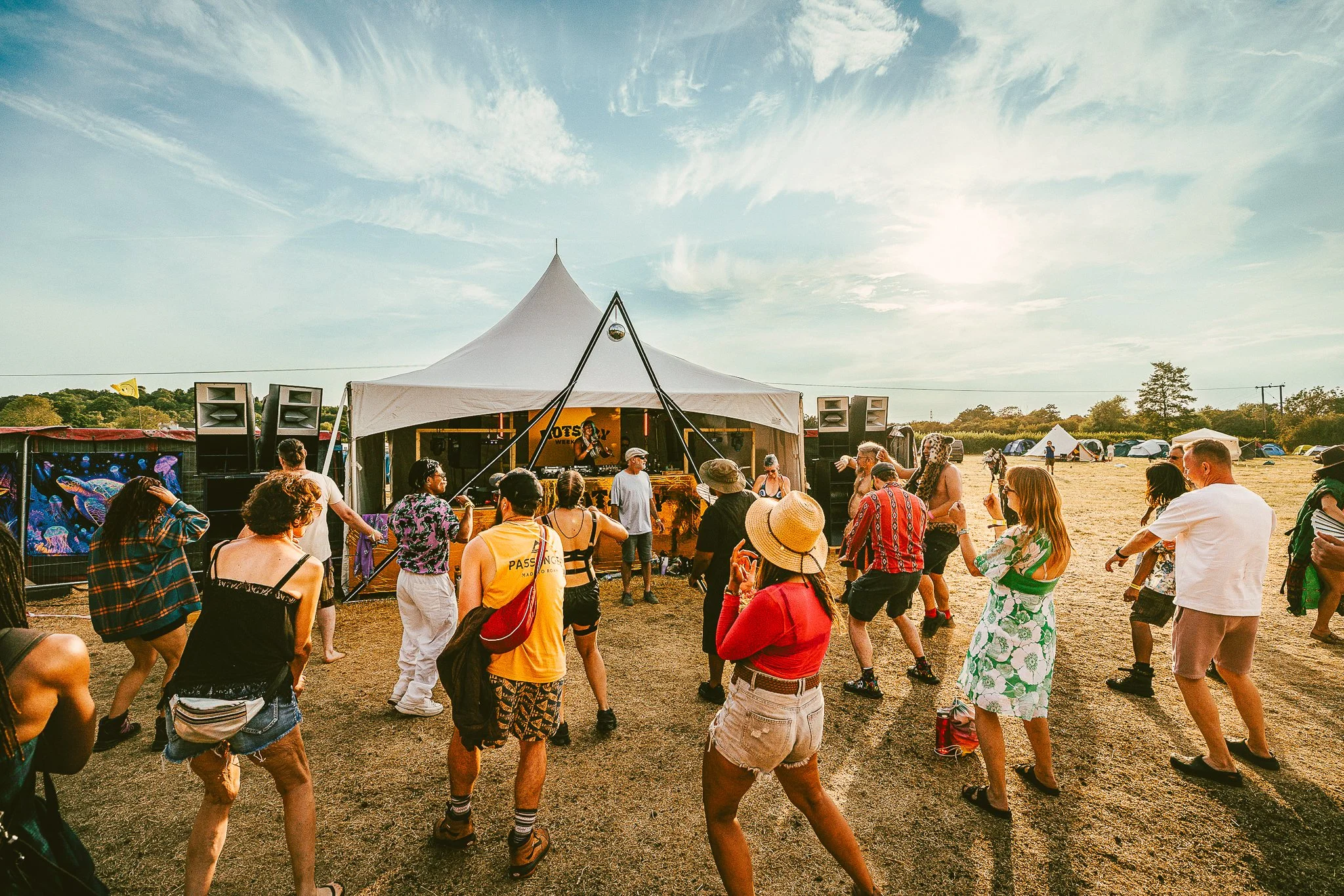 People dancing and enjoying music at an outdoor festival or concert under a large white tent, with a clear sky and scattered clouds in the background.