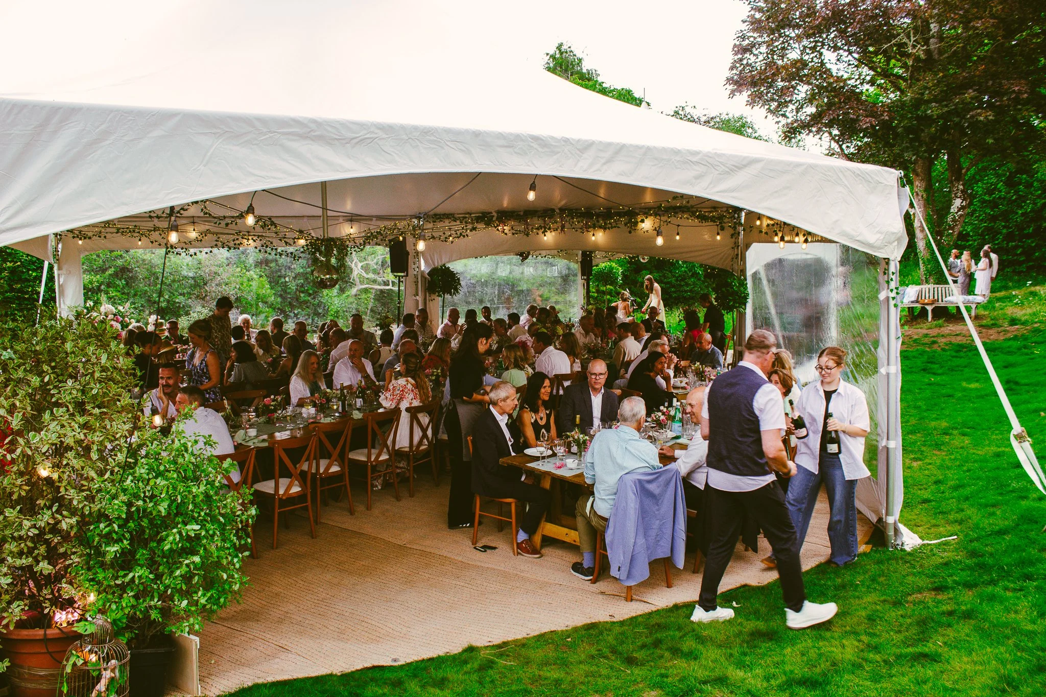 A large outdoor wedding reception under white tent with guests seated at decorated tables, surrounded by greenery and string lights.