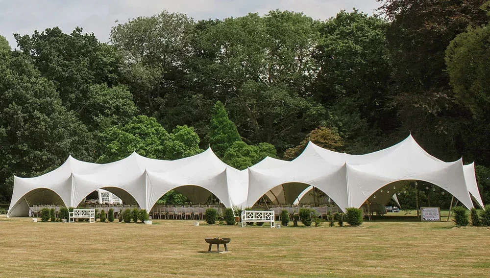 Large white event tent with pointed peaks set up on a grassy field, surrounded by bushes and trees.