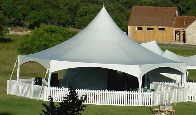 A large white outdoor event tent with a peaked roof, surrounded by a white picket fence, set up on a grassy area with trees and an unfinished wooden building in the background.