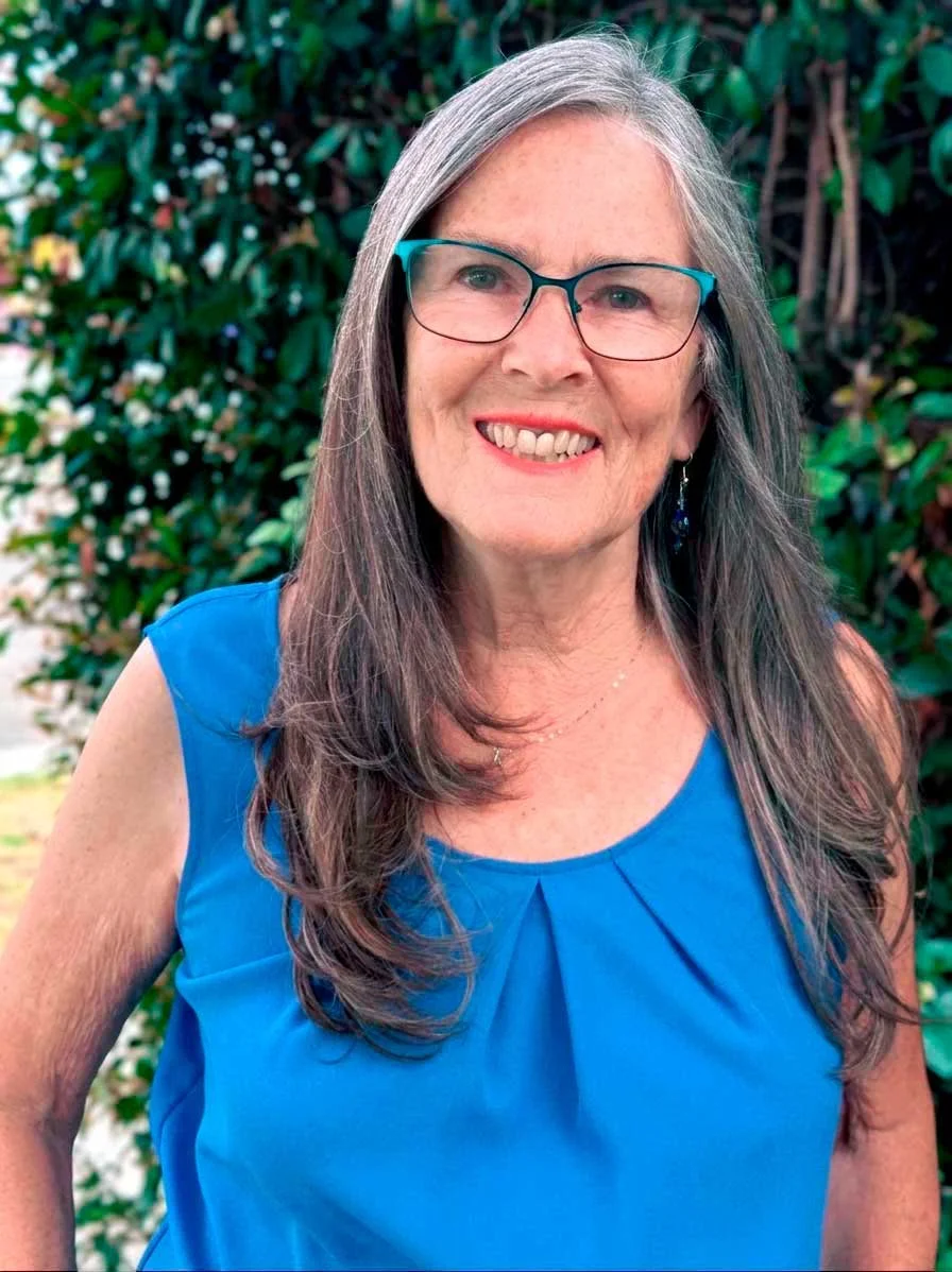 Kaye Main photo of her in front of green bushes. she has a blue shirt on and is happy