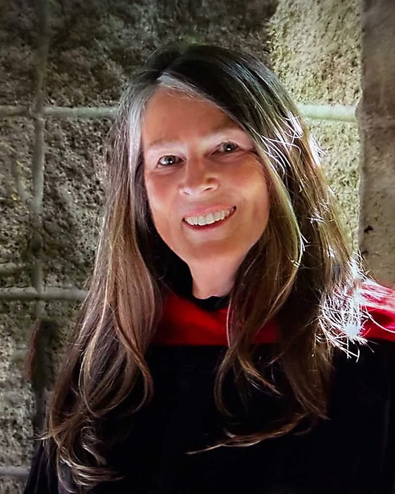 Smiling woman with long brown hair and fair skin, standing against a textured stone wall.