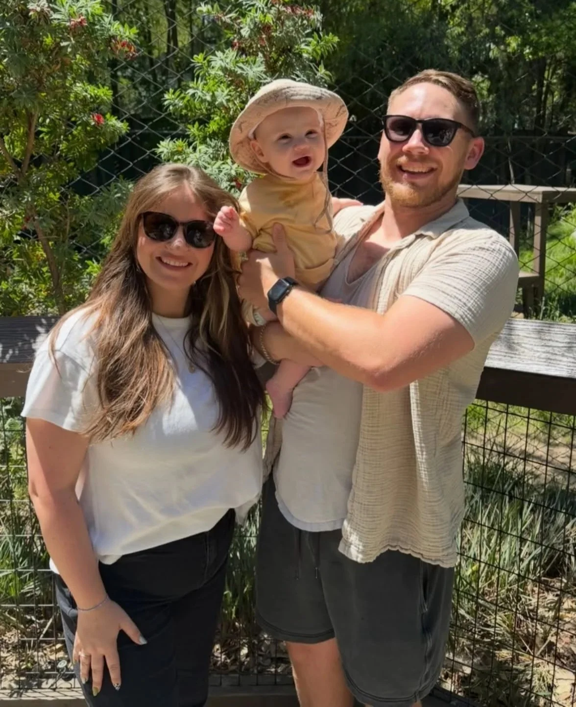 A happy family photo of three people outdoors on a sunny day. A woman with long brown hair and sunglasses stands on the left, a man with short hair, beard, sunglasses, wearing a beige shirt and shorts, holds a baby in a yellow outfit and hat on the right. The background features green trees and a fence.