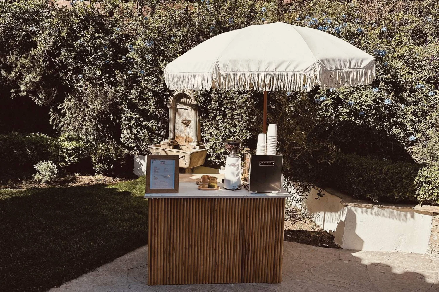 Fluted wood wedding coffee bar beneath a white fringed parasol in a garden courtyard