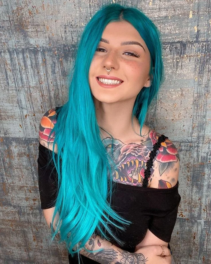 A young woman with long, bright blue hair, freckles, and a septum piercing, smiling in front of a textured, gray background. She is wearing a black off-the-shoulder top that reveals colorful tattoos on her shoulder, chest, and arms.