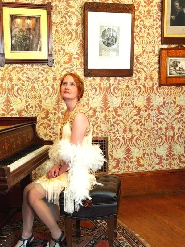 A woman with red hair sitting on a black piano bench in a vintage room with patterned wallpaper, wearing a cream-colored dress with feathered details and black shoes.