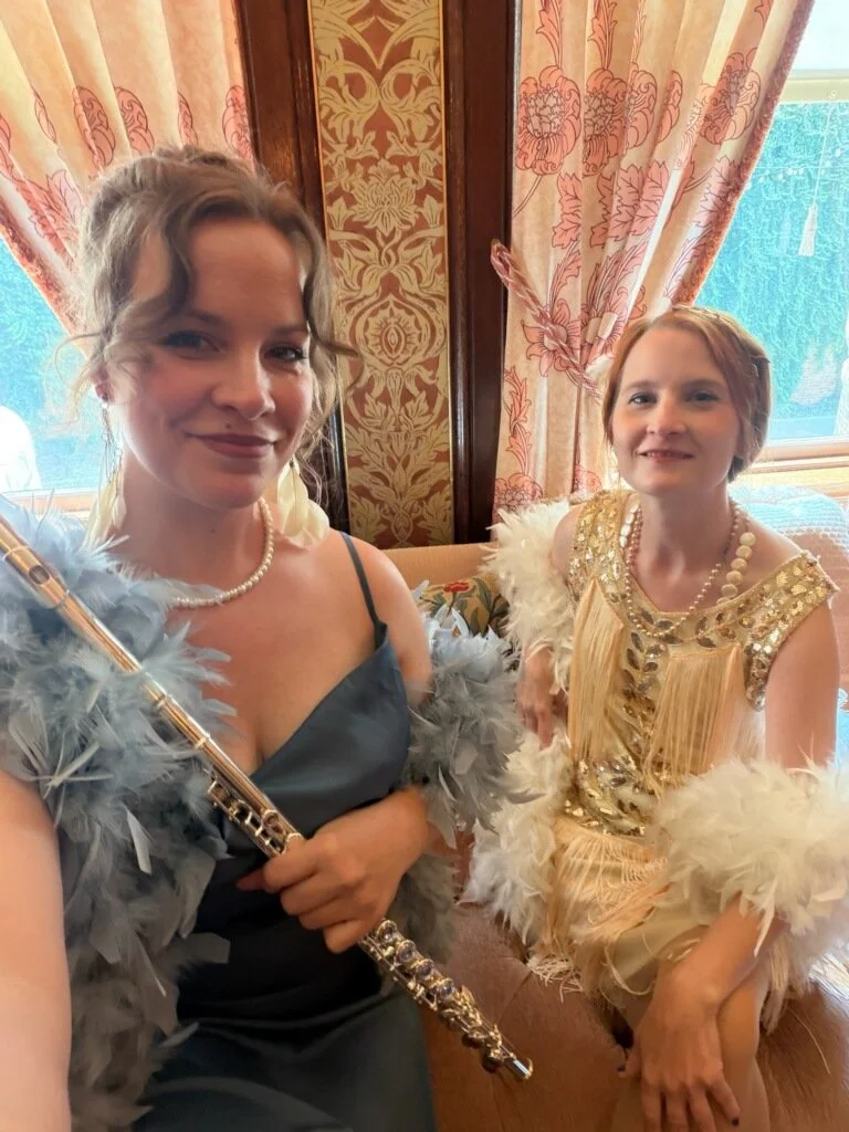 Emily and Caylee dressed in vintage-style costumes with feathered accessories, one holding a flute, sitting in a room with patterned curtains and ornate wallpaper.