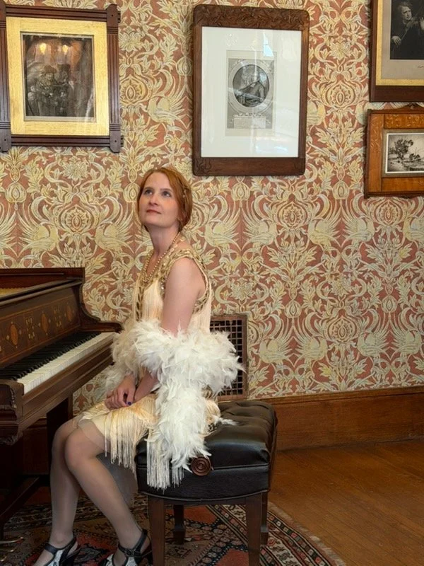 Emily with red hair sitting on a piano bench, dressed in vintage clothing with a feathered shawl, in a room with ornate wallpaper and framed artwork.