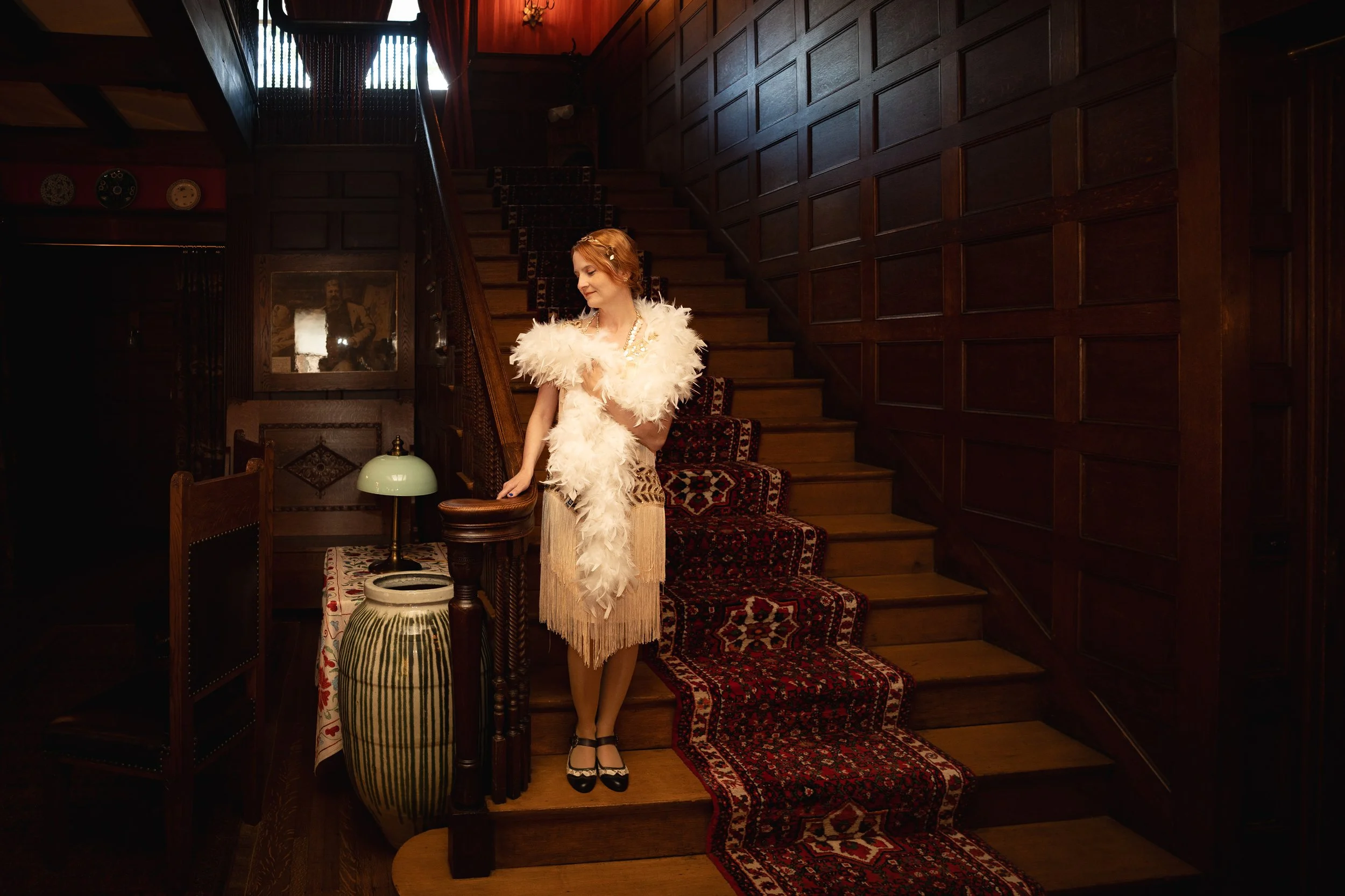 A woman with red hair styled in loose waves, wearing a vintage dress and a large white feather boa, stands on a staircase in a dimly lit wood-paneled room. The decor includes a patterned carpet, a small table with a green lampshade, and framed artwork on the walls.