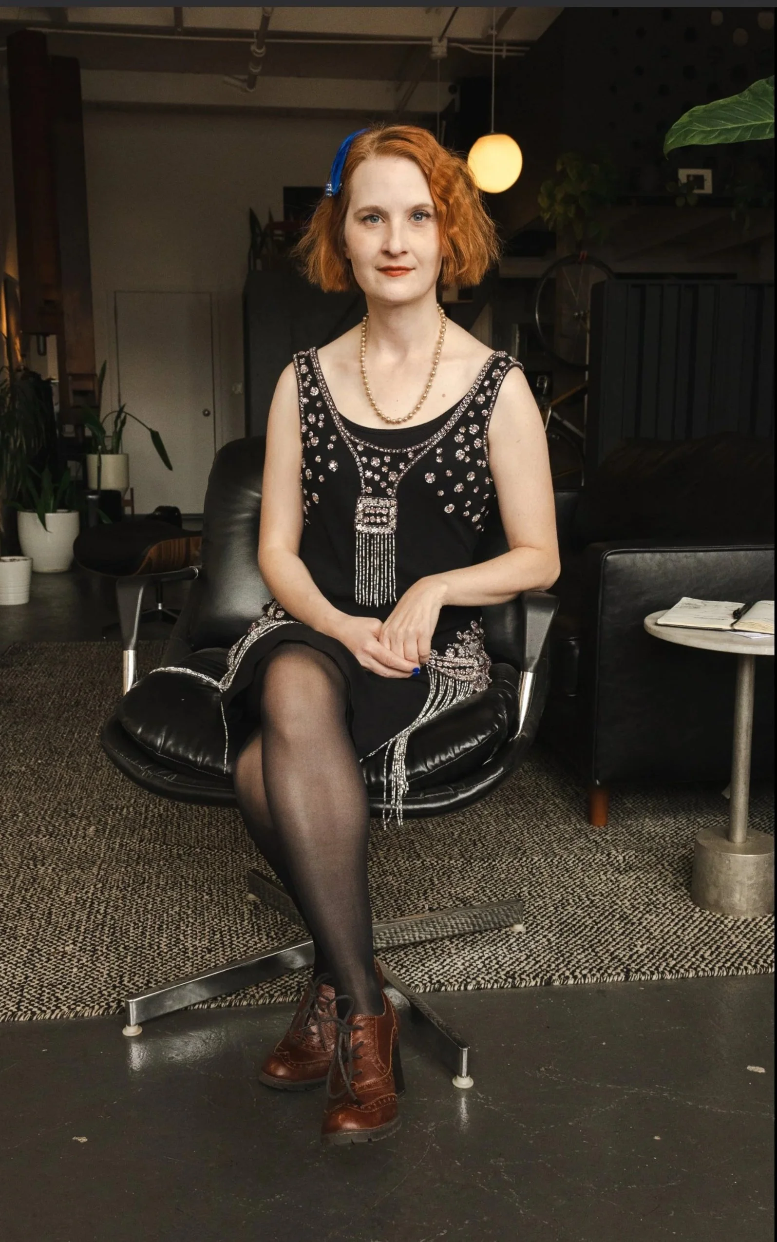 A woman with short red hair, wearing a black dress with rhinestones, black stockings, and brown boots, sitting in a black chair in a modern, artsy indoor space.