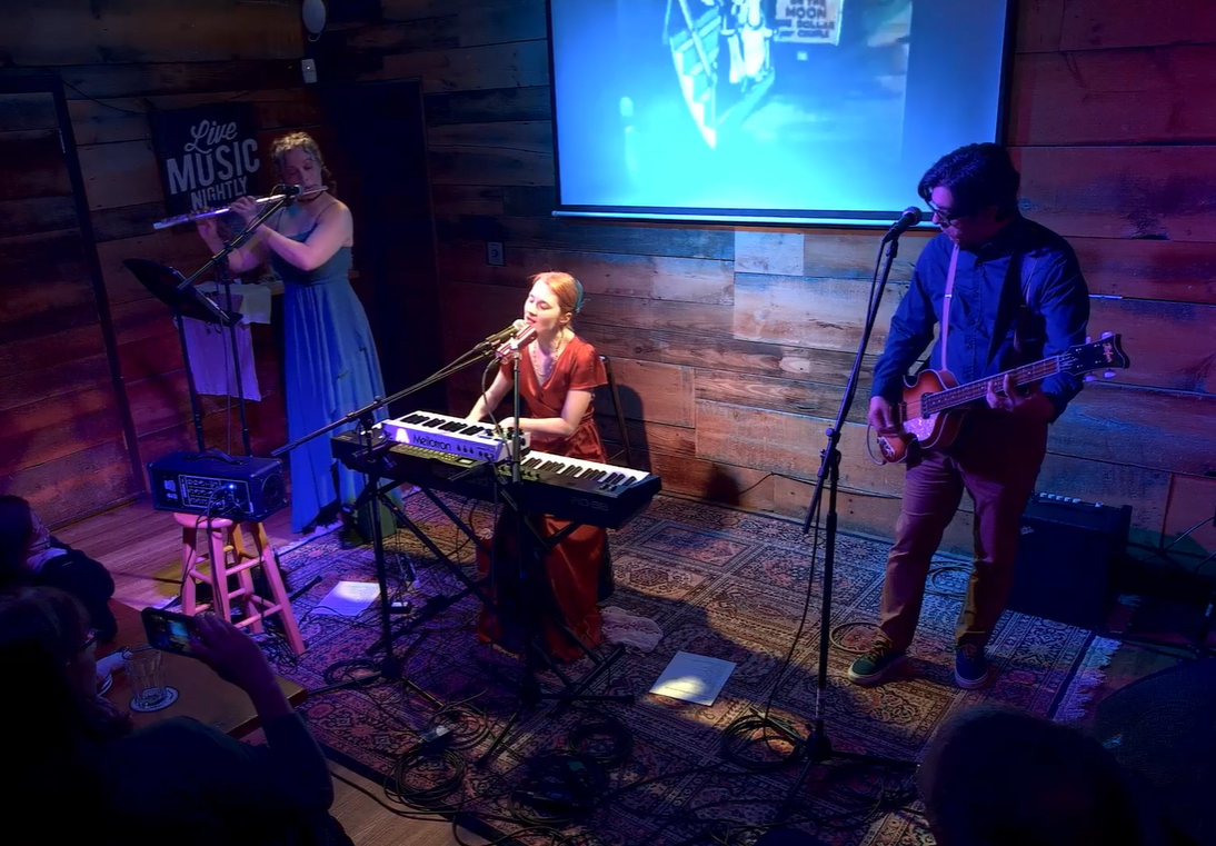 The Emily Chloe Quinn Ensemble performing on a small stage in a wooden venue. Caylee plays the flute, Emily sings and plays a keyboard, and Paul plays an electric guitar. Audience members are visible, with one person taking a photo.